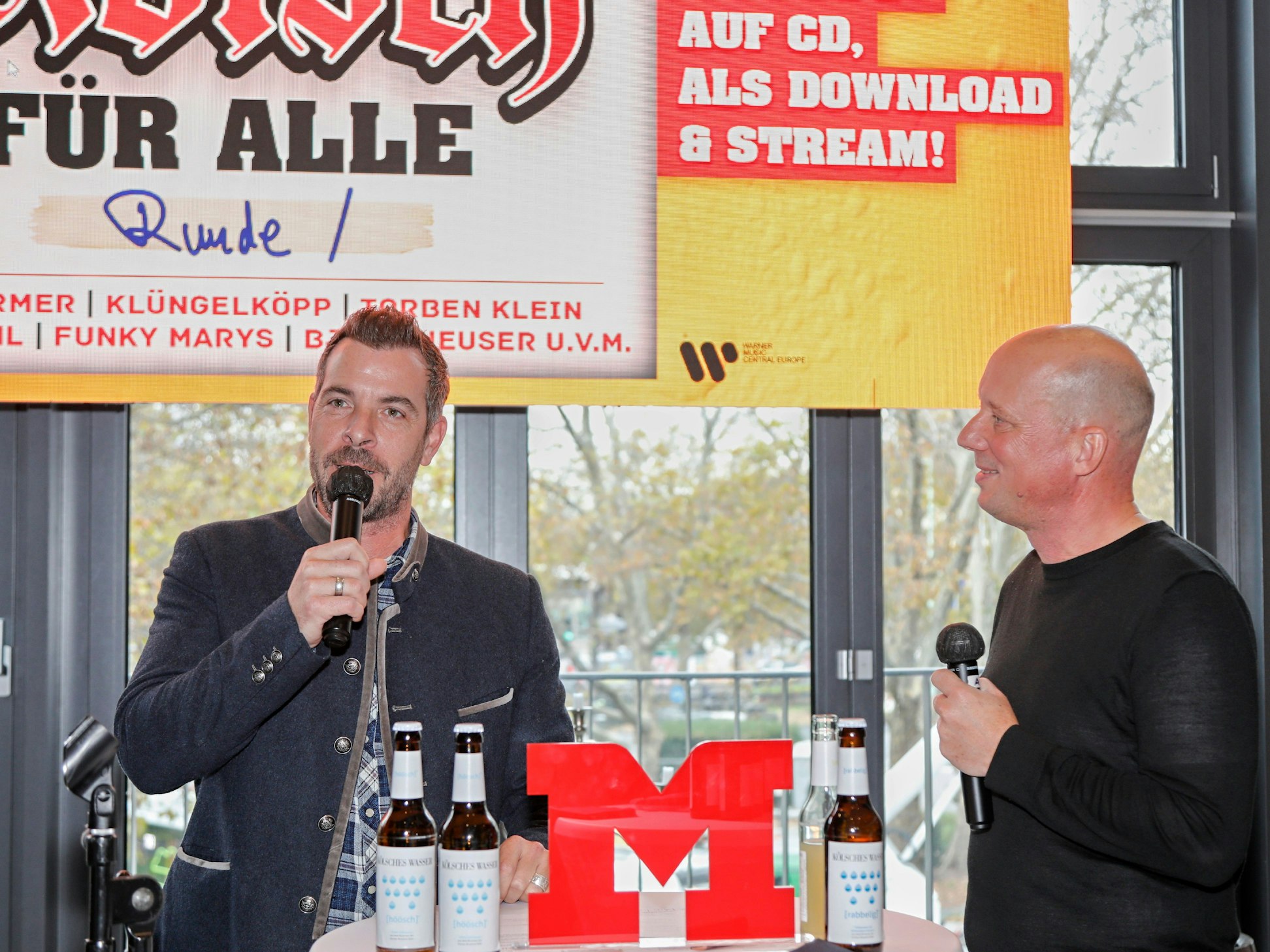 Zwei Männer mit Mikrofon im Gespräch vor einem großen Plakat.