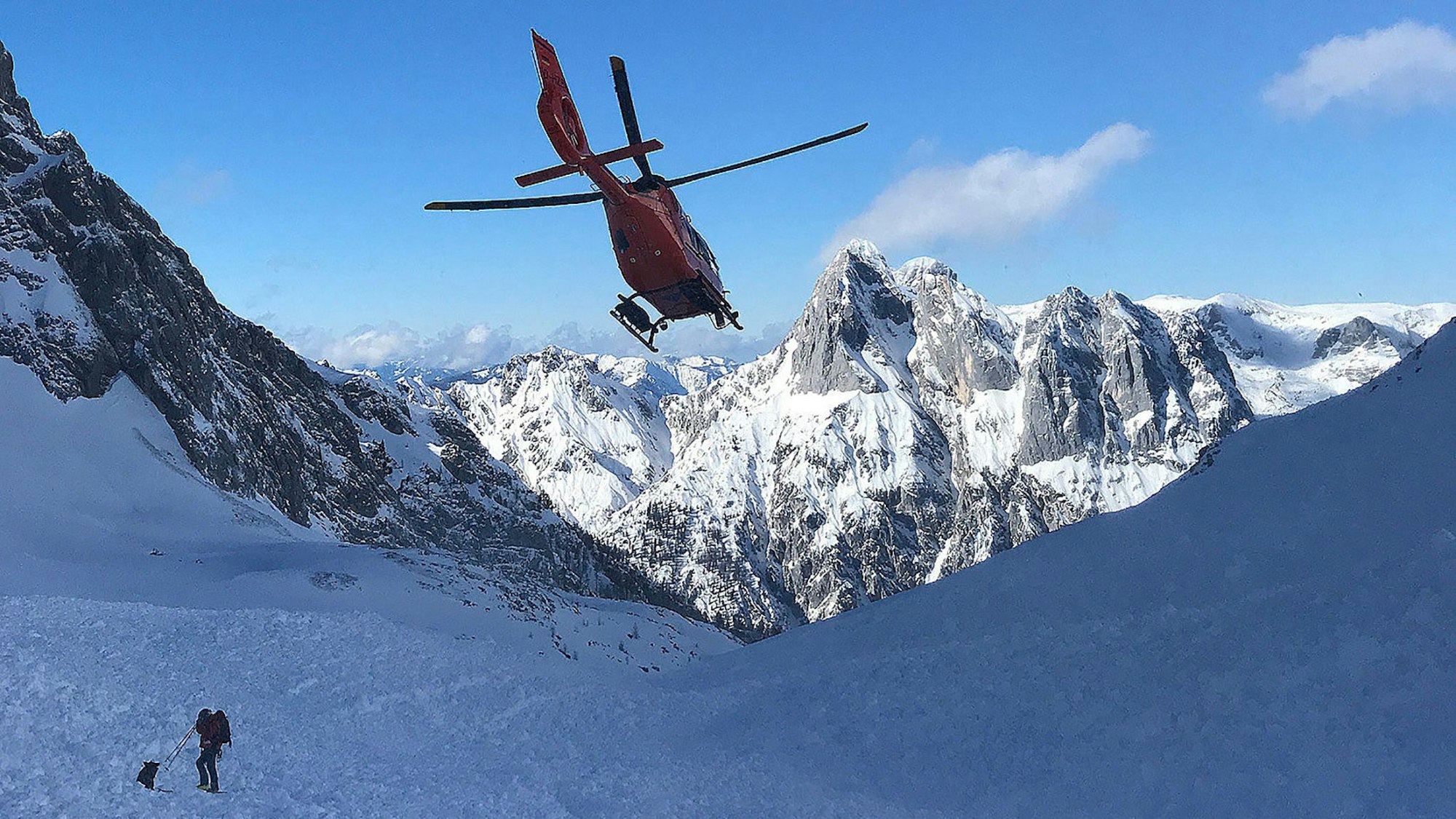 Ein Rettungshubschrauber fliegt über einem Lawinenfeld im Hochkalter-Gebiet (Berchtesgadener Alpen), auf dem ein Retter der Bergwacht mit einem Lawinenhund steht.