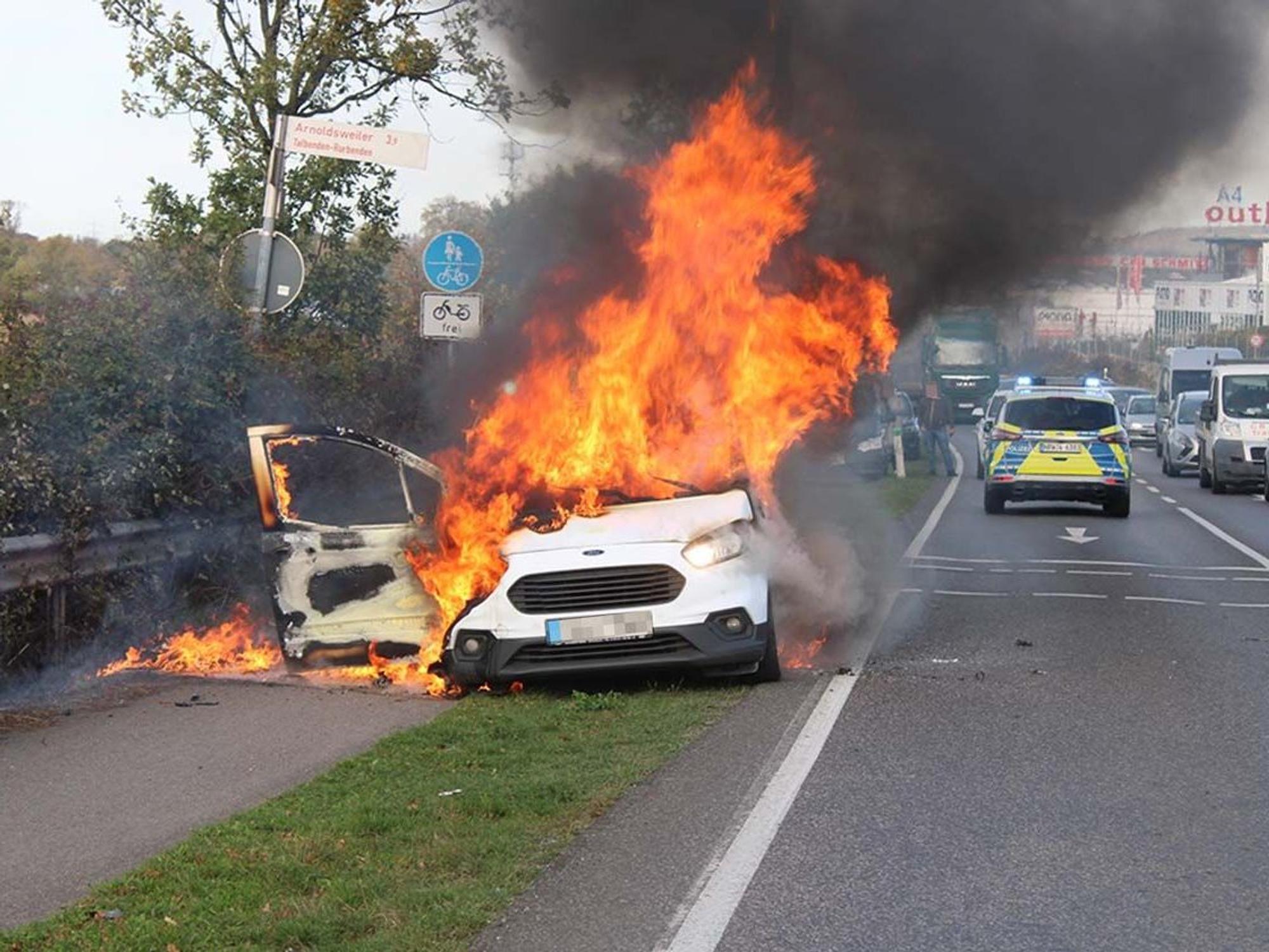 Hohe Flammen schlagen aus einem Auto, das direkt an einer Straße steht.