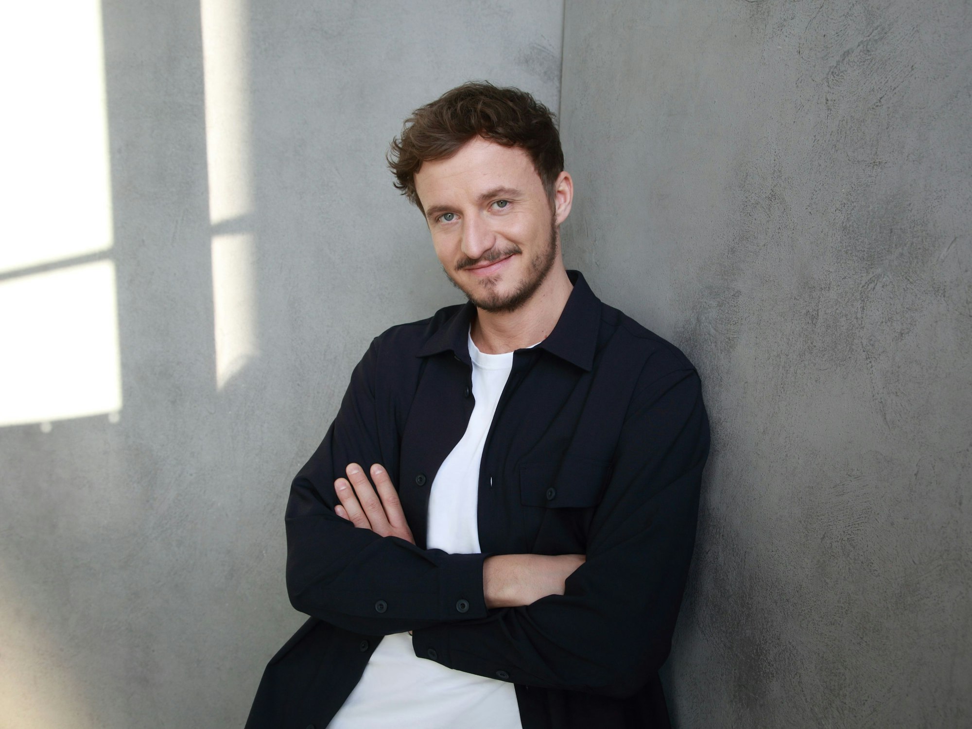 Moderator Tommi Schmitt lehnt an einer Wand und lächelt in die Kamera.