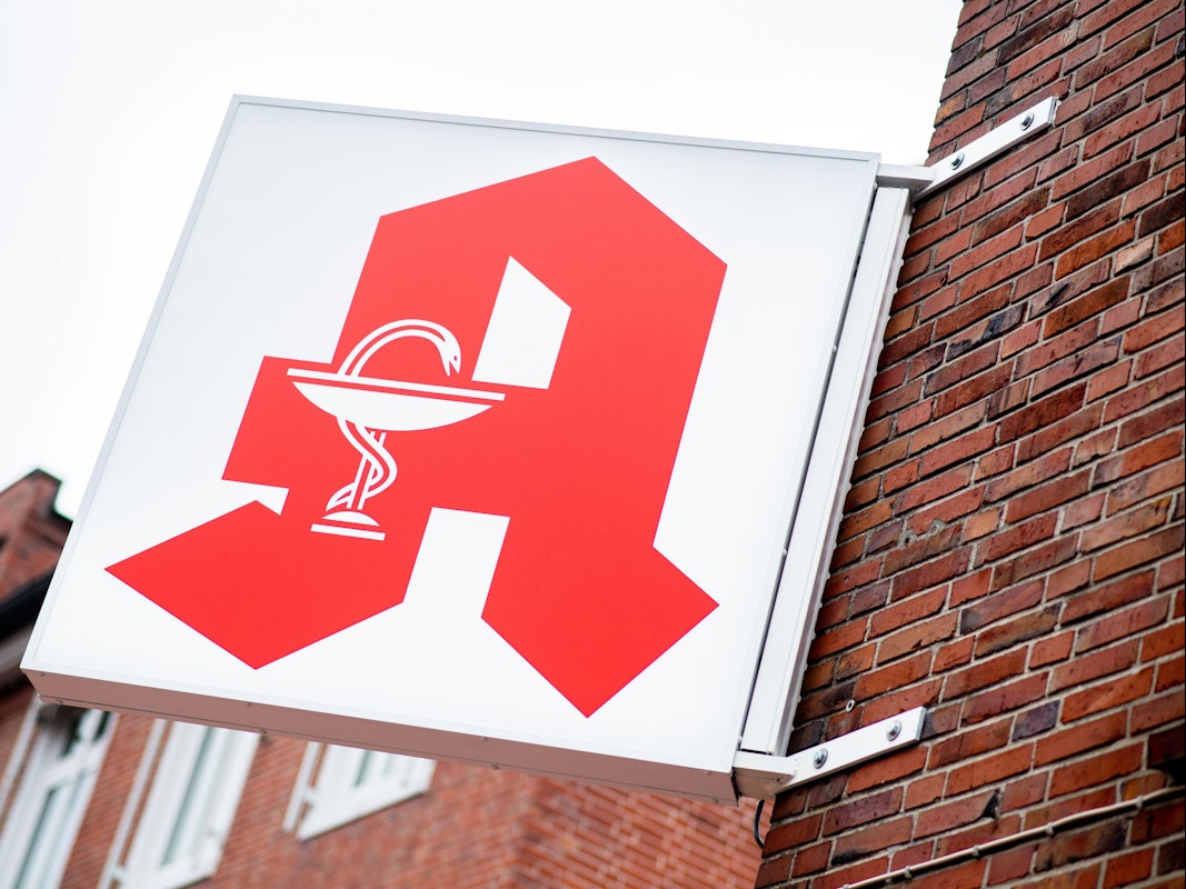 Das Schild für öffentliche Apotheken hängt an einer Hauswand.