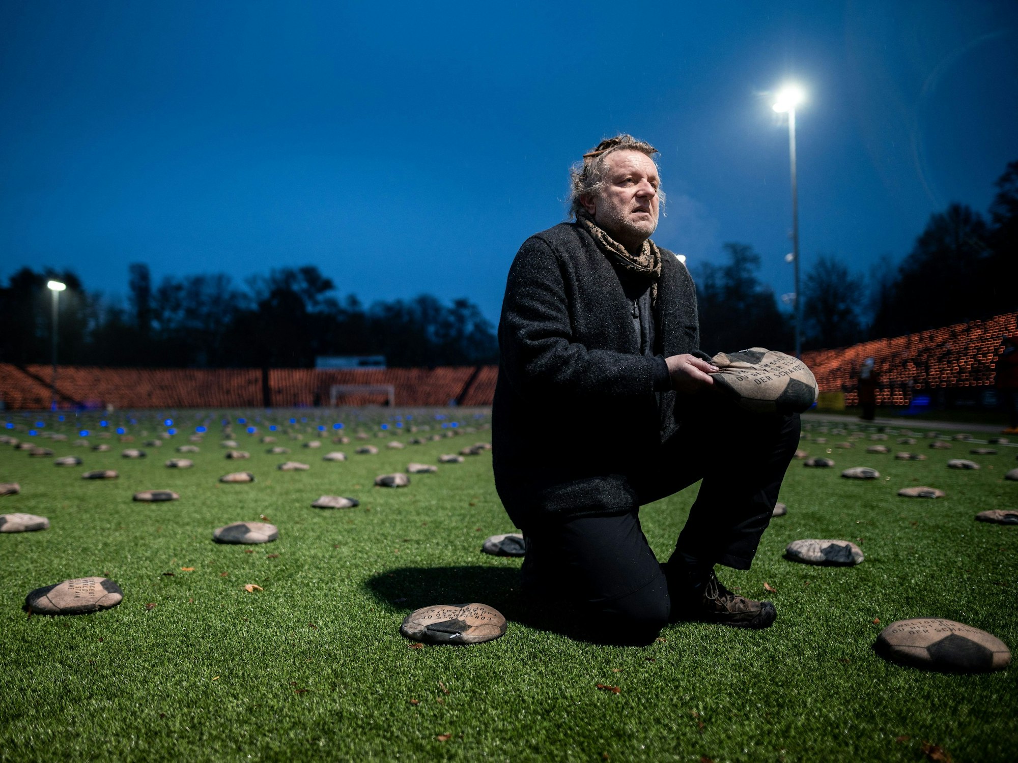 Der Künstler Volker-Johannes Trieb kniet auf dem Rasen und hält einen Stoff-Fußball in der Hand.