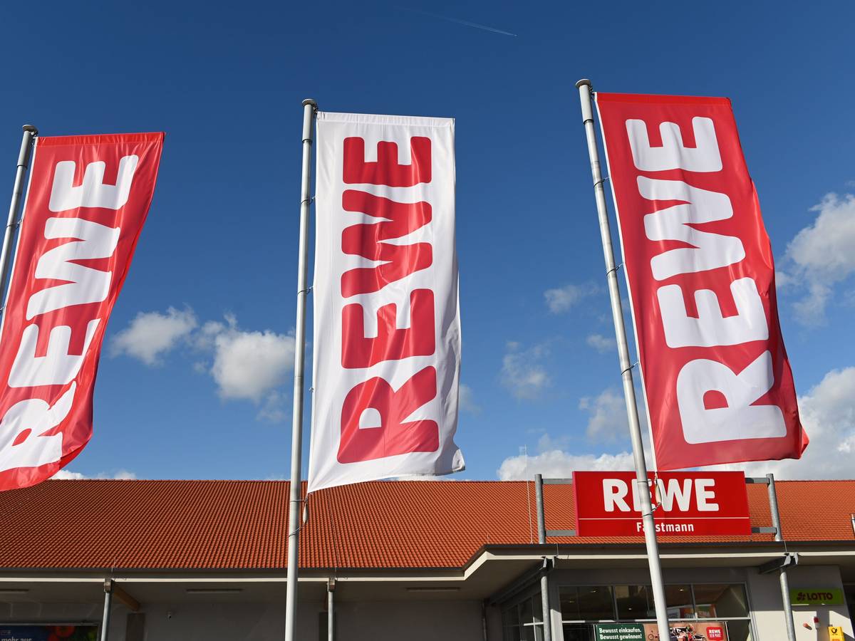 Fahnen mit dem Logo des Lebensmitteleinzelhändlers Rewe wehen vor einer Filiale.