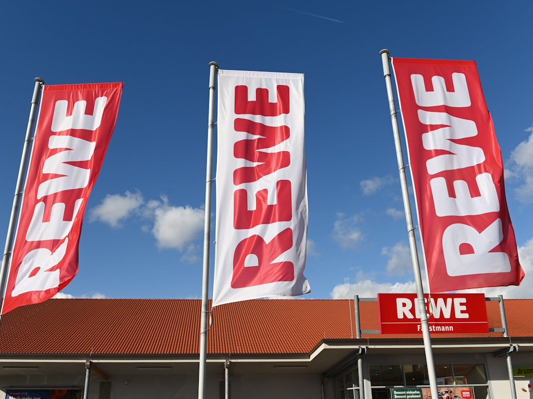 Fahnen mit dem Logo des Lebensmitteleinzelhändlers Rewe wehen vor einer Filiale.