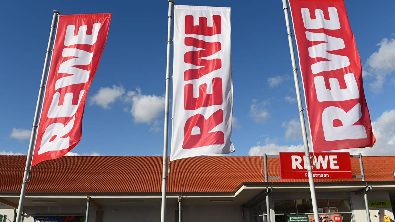 Fahnen mit dem Logo des Lebensmitteleinzelhändlers Rewe wehen vor einer Filiale.