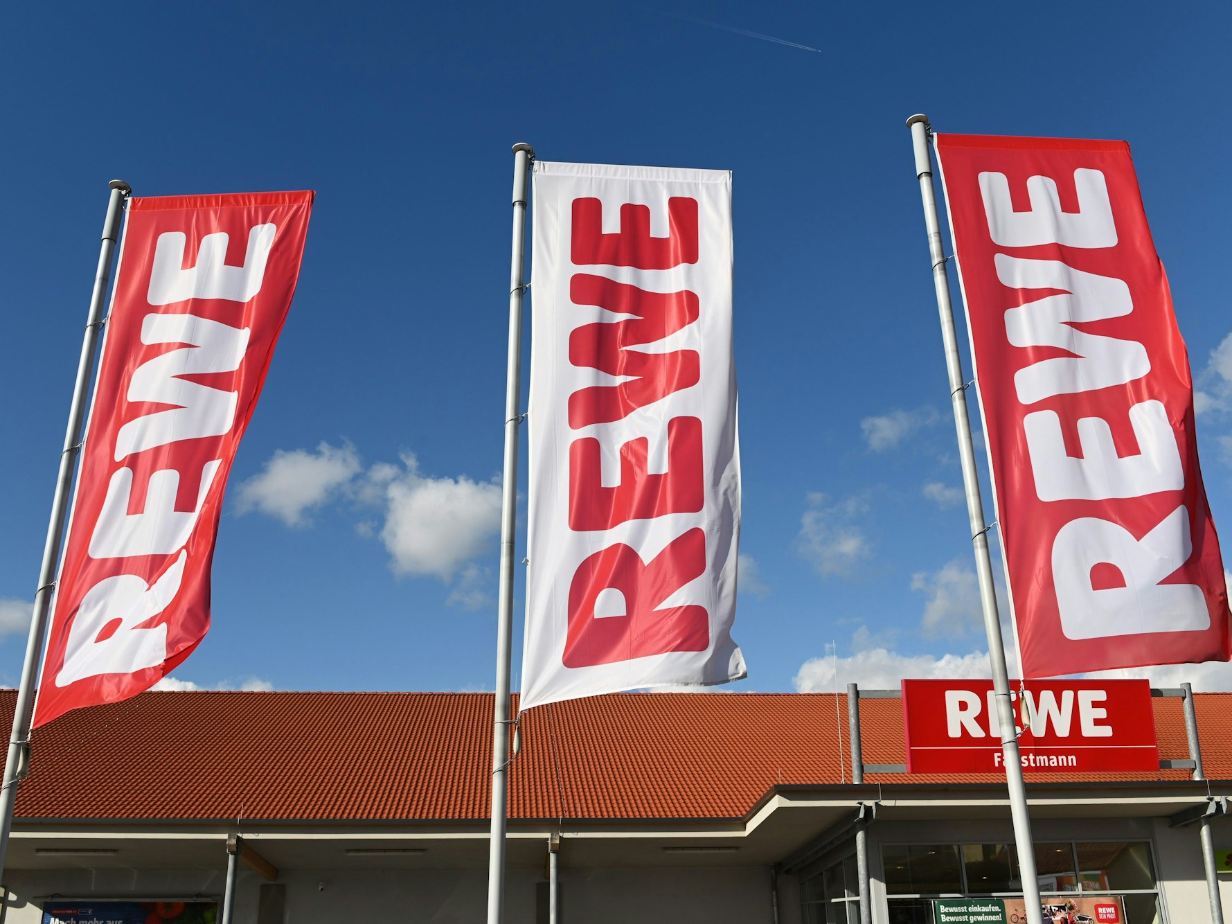 Fahnen mit dem Logo des Lebensmitteleinzelhändlers Rewe wehen vor einer Filiale.