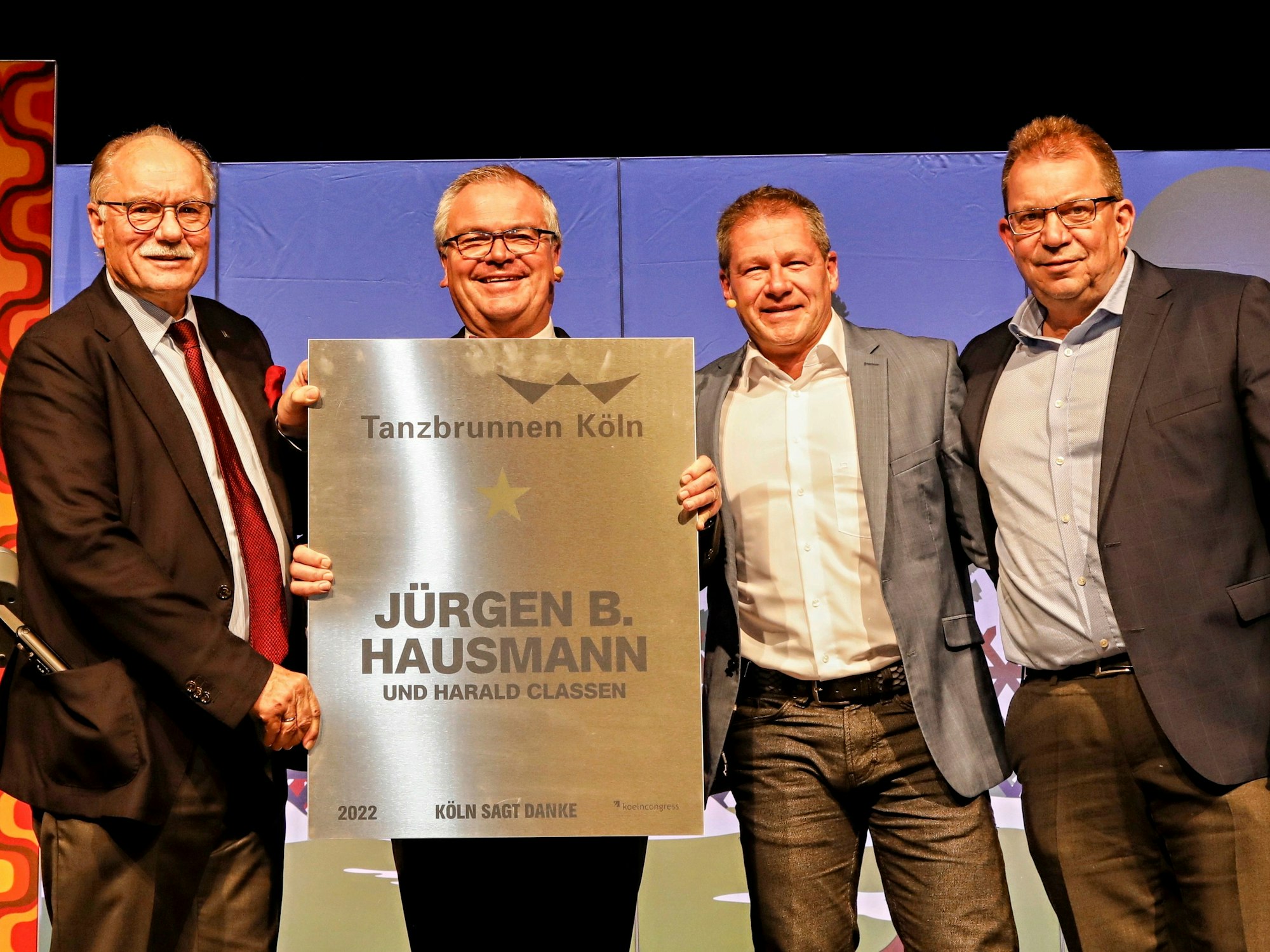 Vier Männer stehen auf einer Bühne und zeigen grinsend eine goldene Ehrentafel in die Kamera.