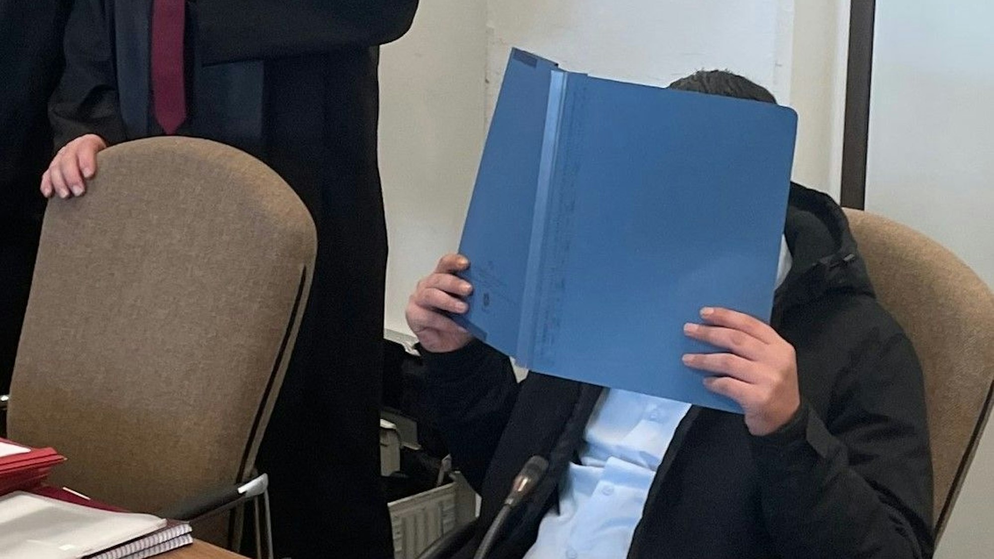Der Angeklagte hält sich einen Ordner vor das Gesicht.