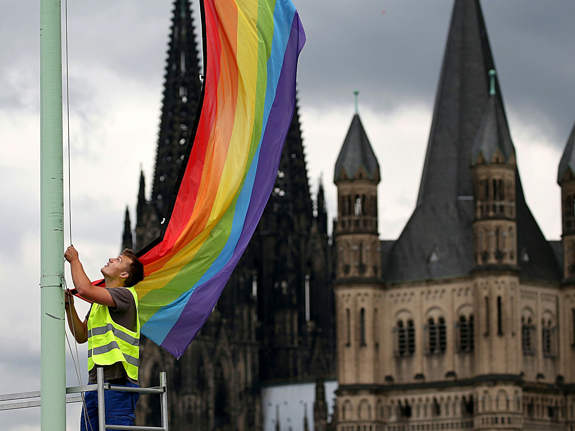 Ein Arbeiter hängt in Köln vor der Kulisse des Doms und der Kirche Groß St. Martin eine Regenbogenfahne auf.