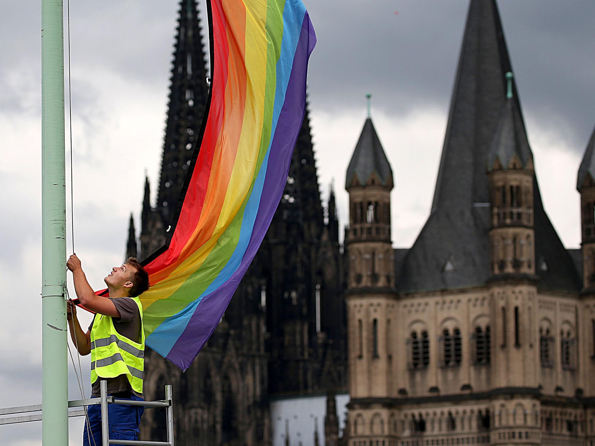 Ein Arbeiter hängt in Köln vor der Kulisse des Doms und der Kirche Groß St. Martin eine Regenbogenfahne auf.