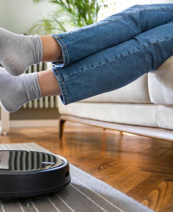 Ein Saugroboter saugt den Boden und eine Frau sitzt auf dem Sofa und hält die Füße hoch, damit das Gerät darunter durchfahren kann.