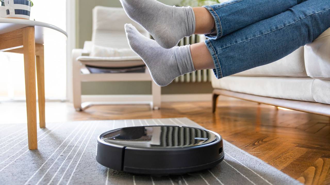 Ein Saugroboter saugt den Boden und eine Frau sitzt auf dem Sofa und hält die Füße hoch, damit das Gerät darunter durchfahren kann.