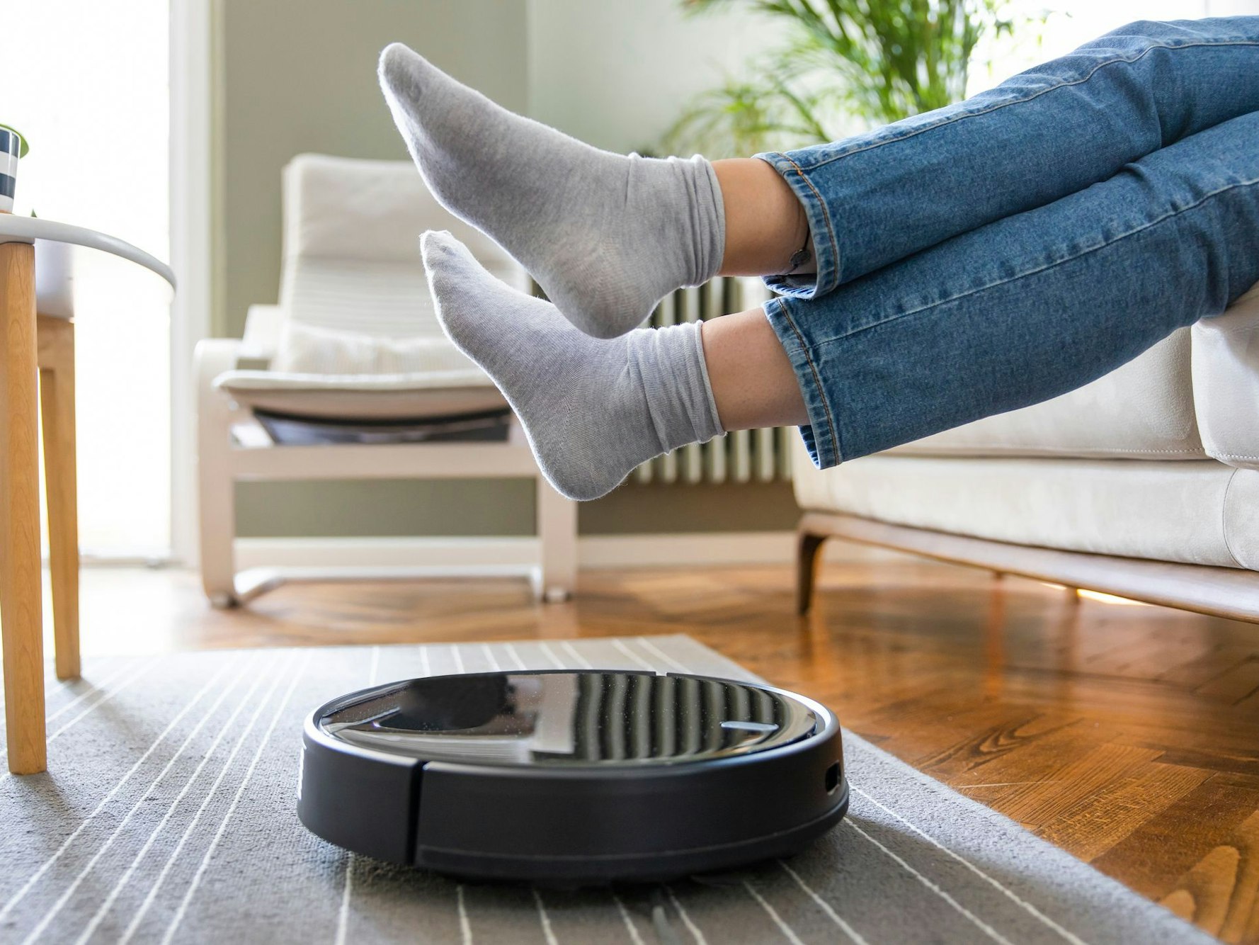 Ein Saugroboter saugt den Boden und eine Frau sitzt auf dem Sofa und hält die Füße hoch, damit das Gerät darunter durchfahren kann.