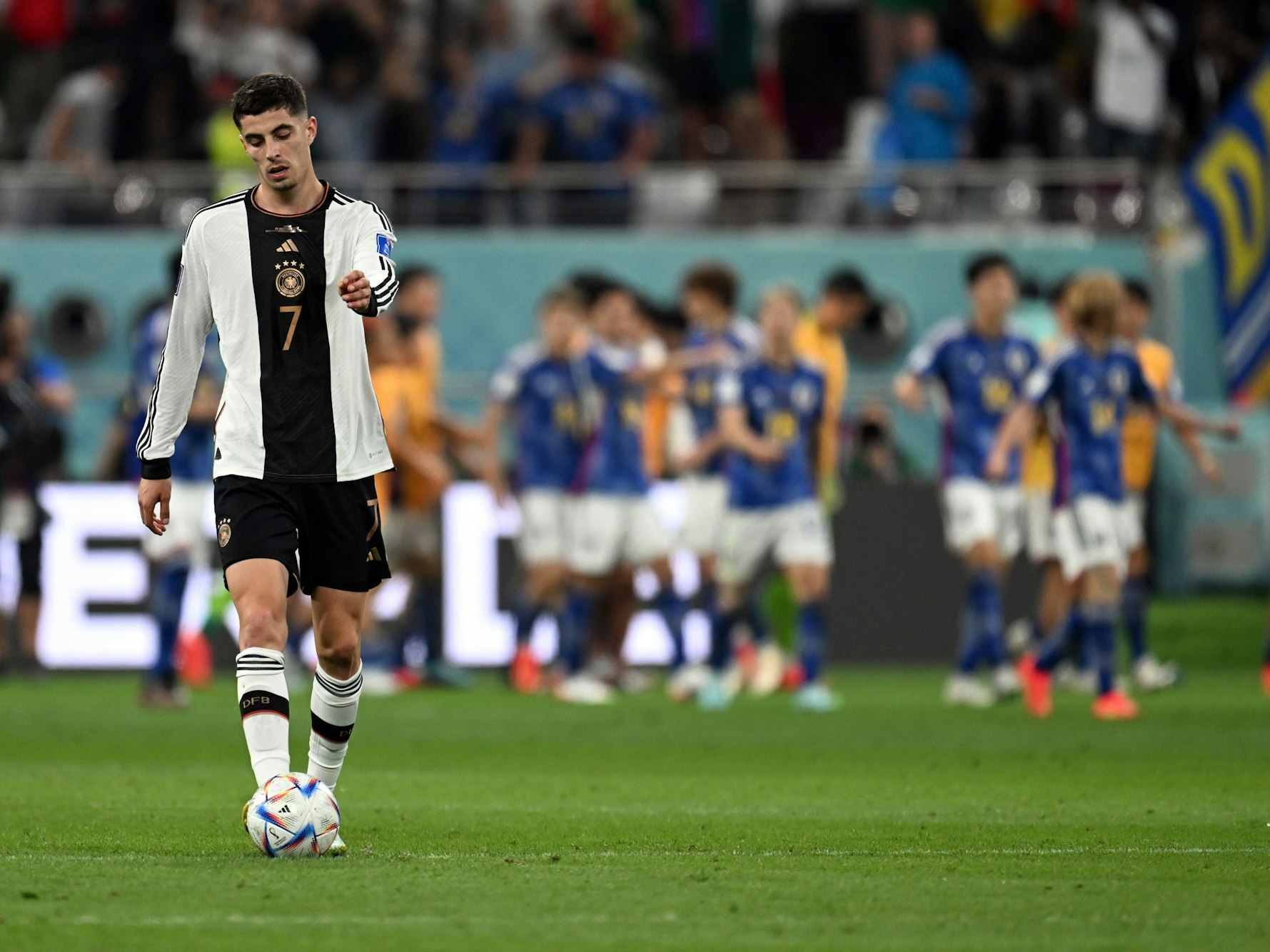 Kai Havertz geht mit gesenktem Kopf über den Platz. Im Hintergrund feiern die siegreichen Japaner.