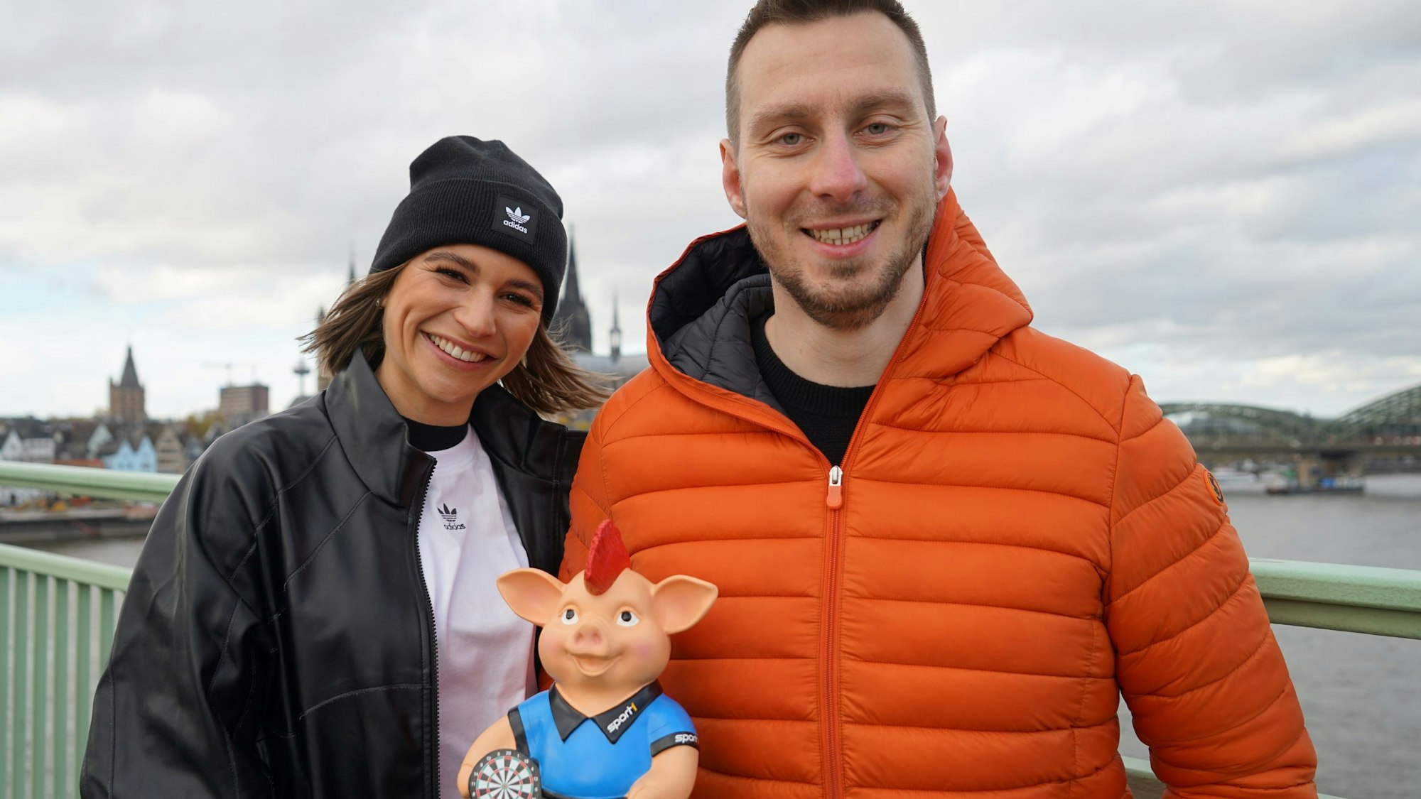 Darts-Profi Florian Hempel und Sport1-Moderatorin Jana Wosnitza lächeln gemeinsam in die Kamera.