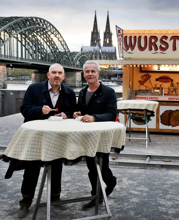 Die beiden Kölner Tatort-Kommissare stehen vor einer Bratwurst-Bude und dem Dom-Panorama.