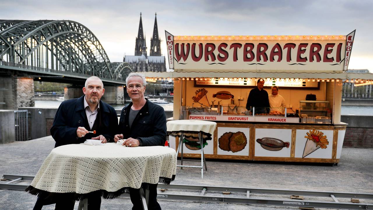 Die beiden Kölner Tatort-Kommissare stehen vor einer Bratwurst-Bude und dem Dom-Panorama.