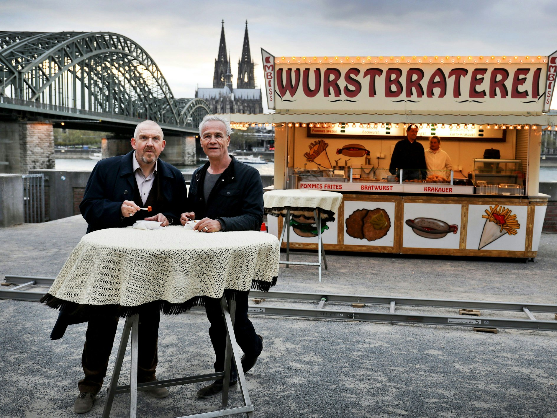 Die beiden Kölner Tatort-Kommissare stehen vor einer Bratwurst-Bude und dem Dom-Panorama.
