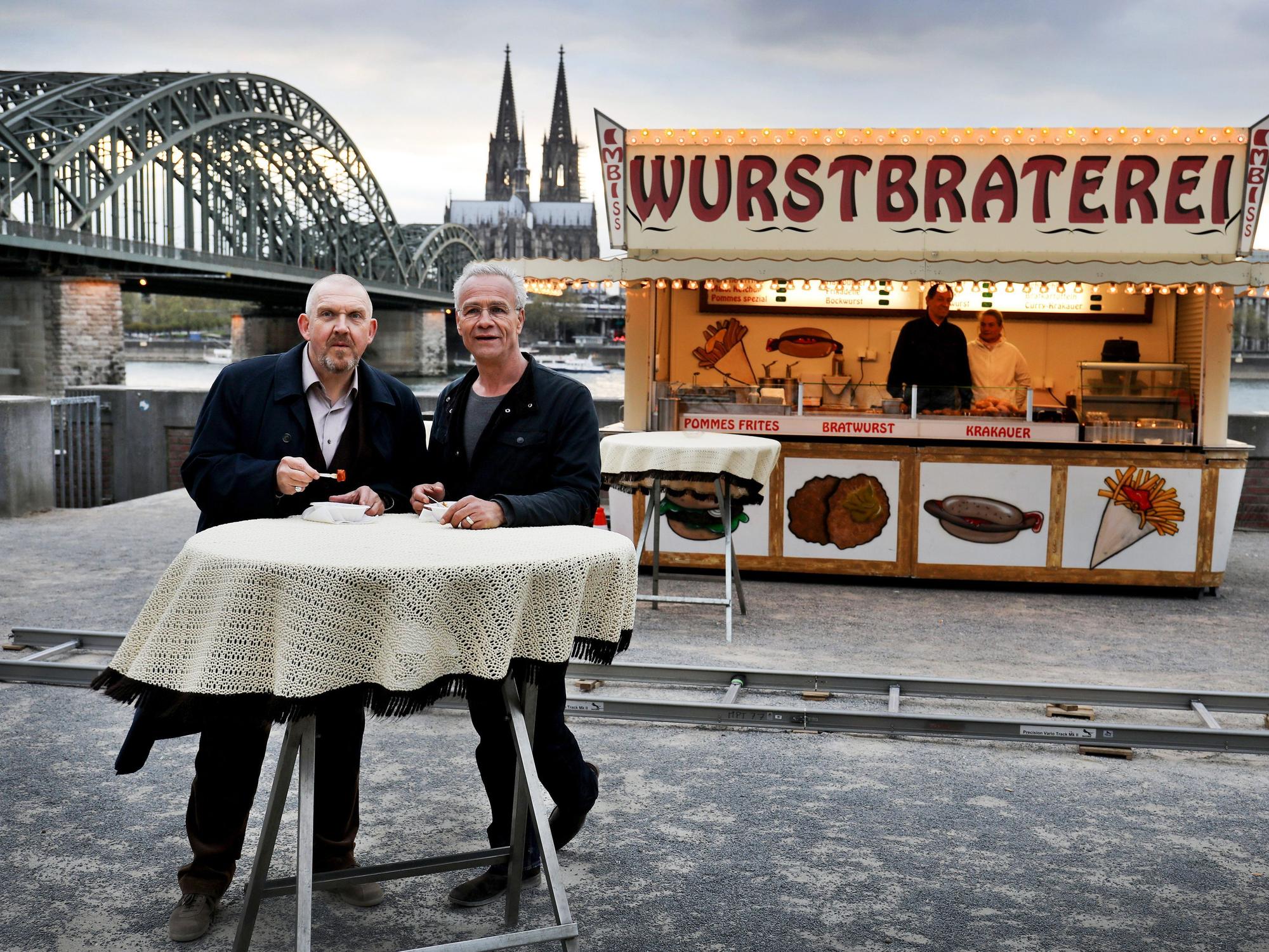 Die beiden Kölner Tatort-Kommissare stehen vor einer Bratwurst-Bude und dem Dom-Panorama.