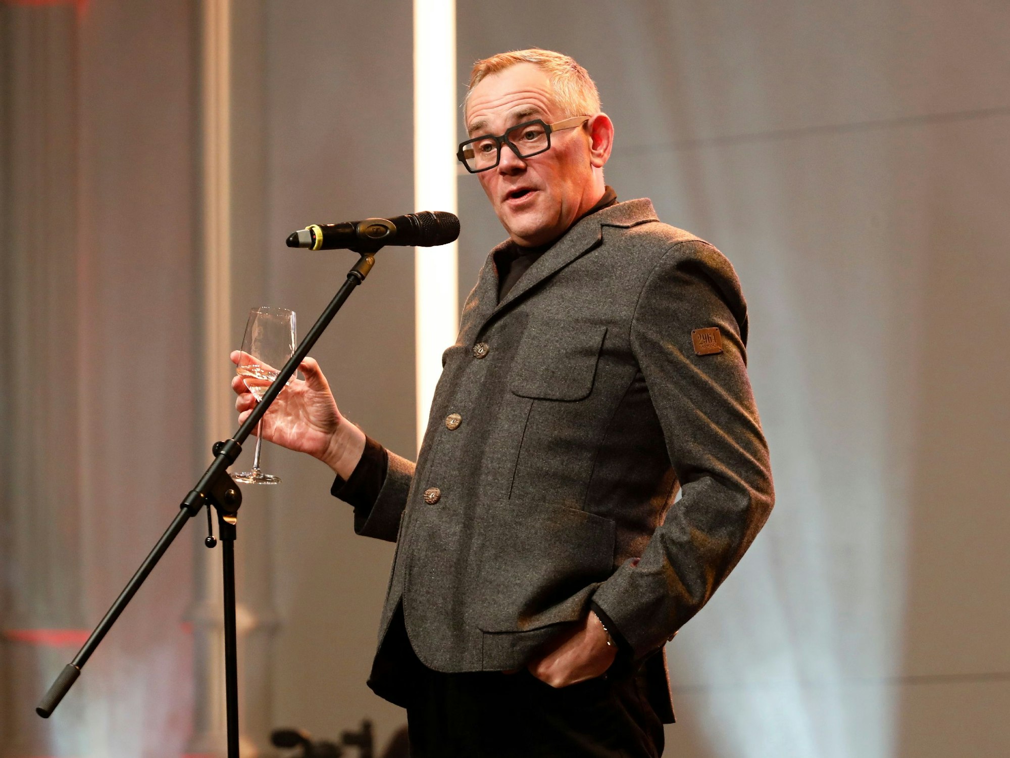 Mann in grauem Anzug steht entspannt mit einem Glas Wein vor einem Mikrofon und spricht mit dem Publikum.