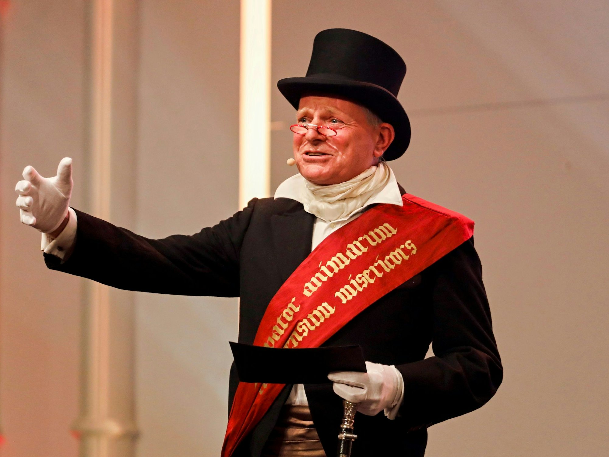 Mann in schwarzem Anzug und mit Hut moderiert eine Sendung. Er liest Text von einer Karte ab.