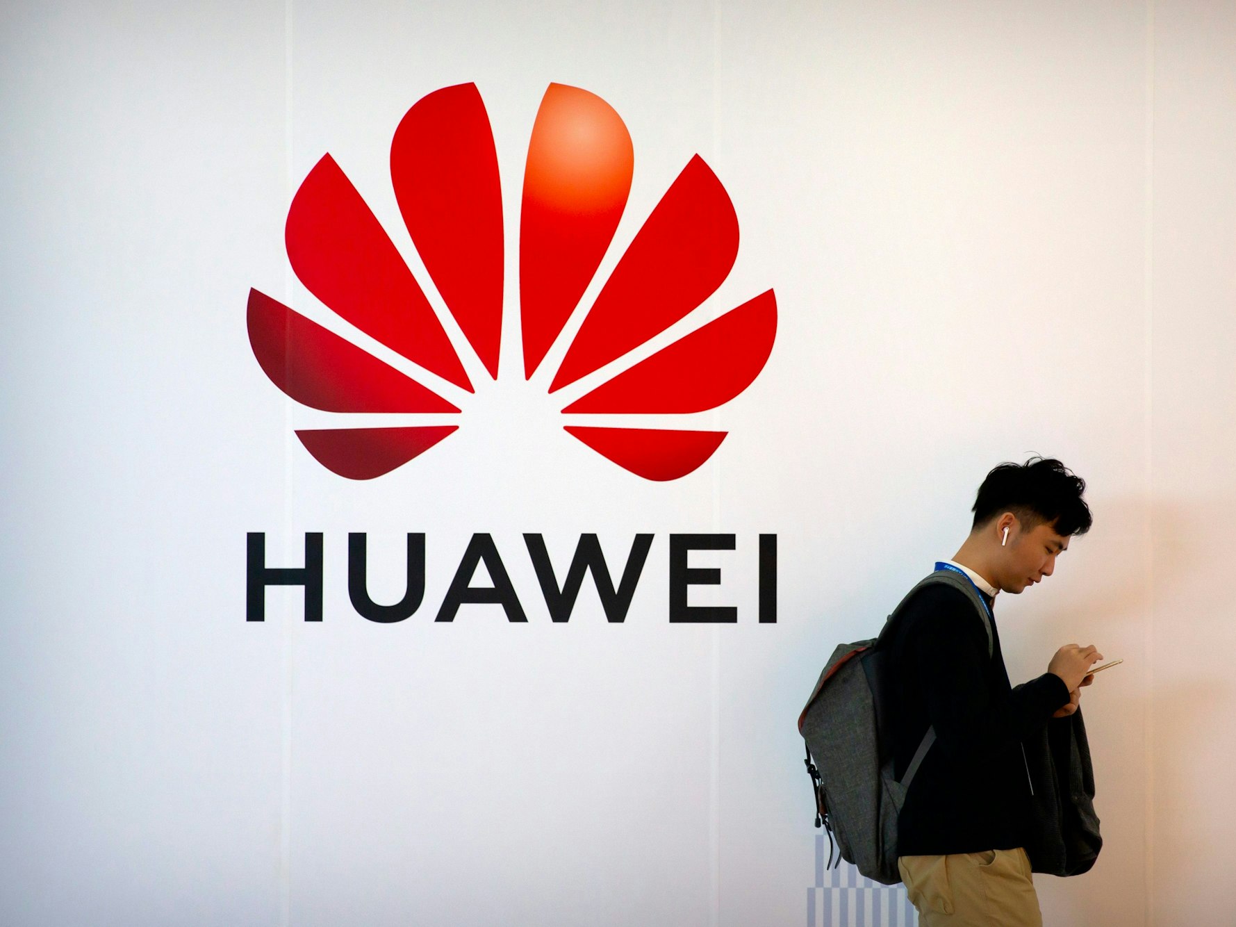 Ein Mann tippt auf seinem Smartphone, während er auf der Fachmesse PT Expo in der Nähe einer Werbetafel für die chinesische Technologiefirma Huawei steht.
