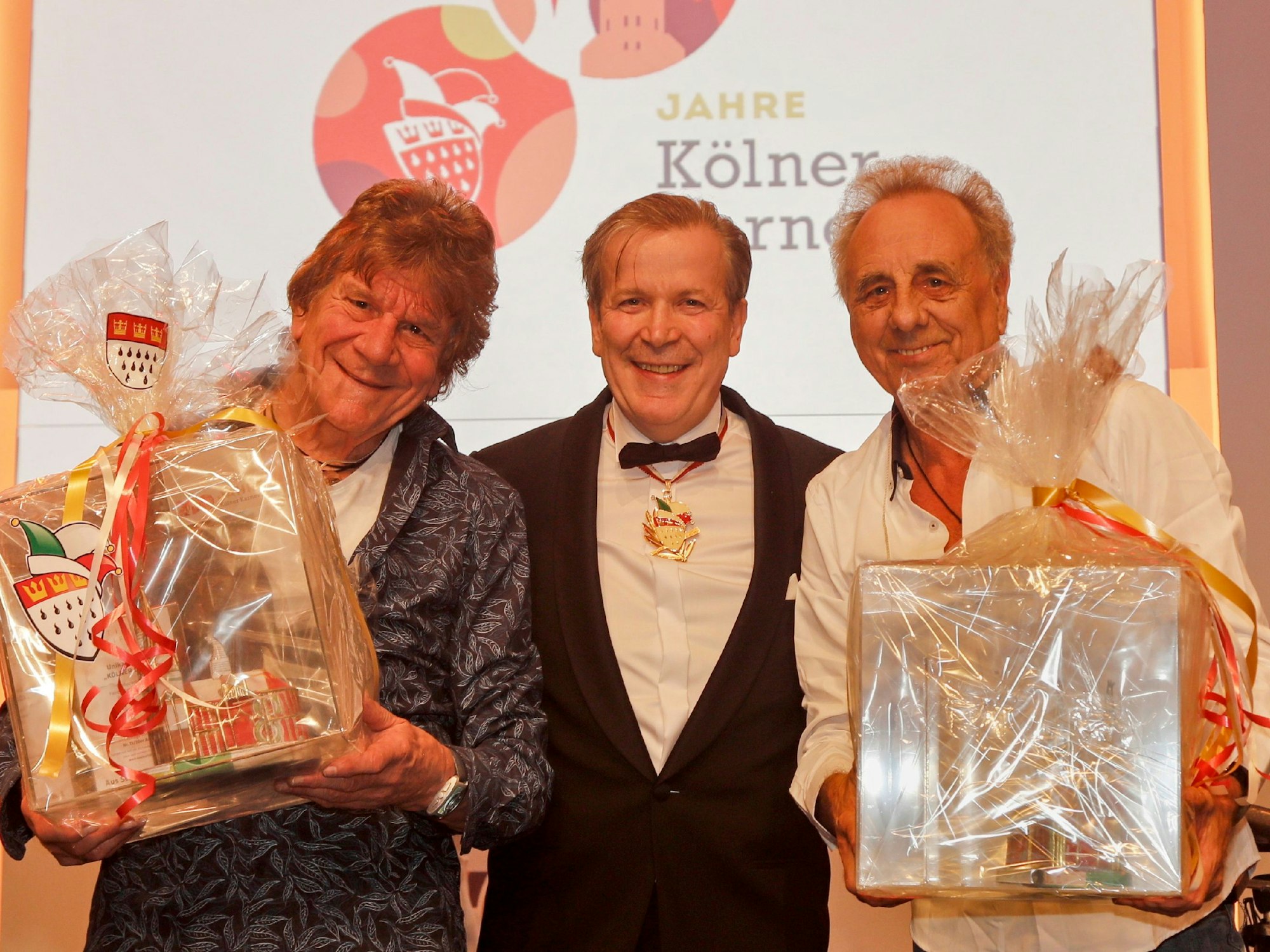 Drei Männer lachen gemeinsam in die Kamera, zwei von ihnen haben Geschenke in der Hand.