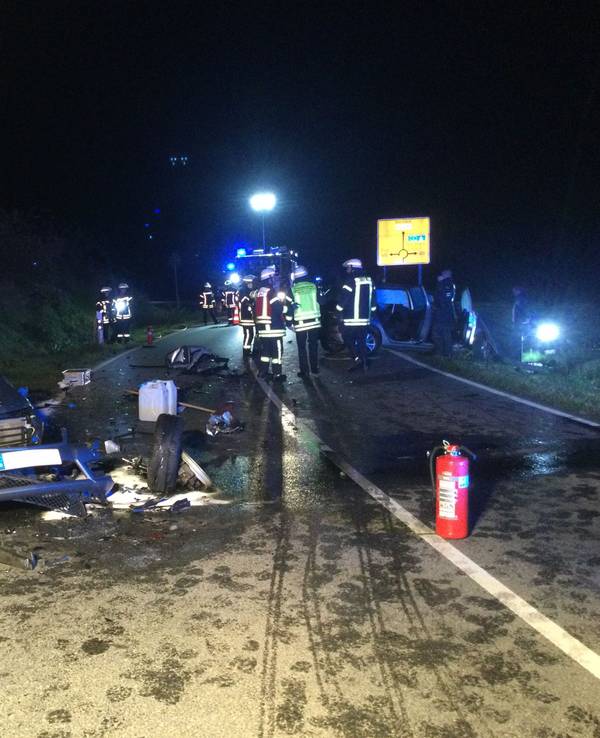 Ein beschädigtes Auto steht schräg neben der Straße, um es herum liegen Trümmerteile. Ein paar Meter weiter befinden sich mehrere Einsatzkräfte der Feuerwehr auf und neben der Fahrbahn.