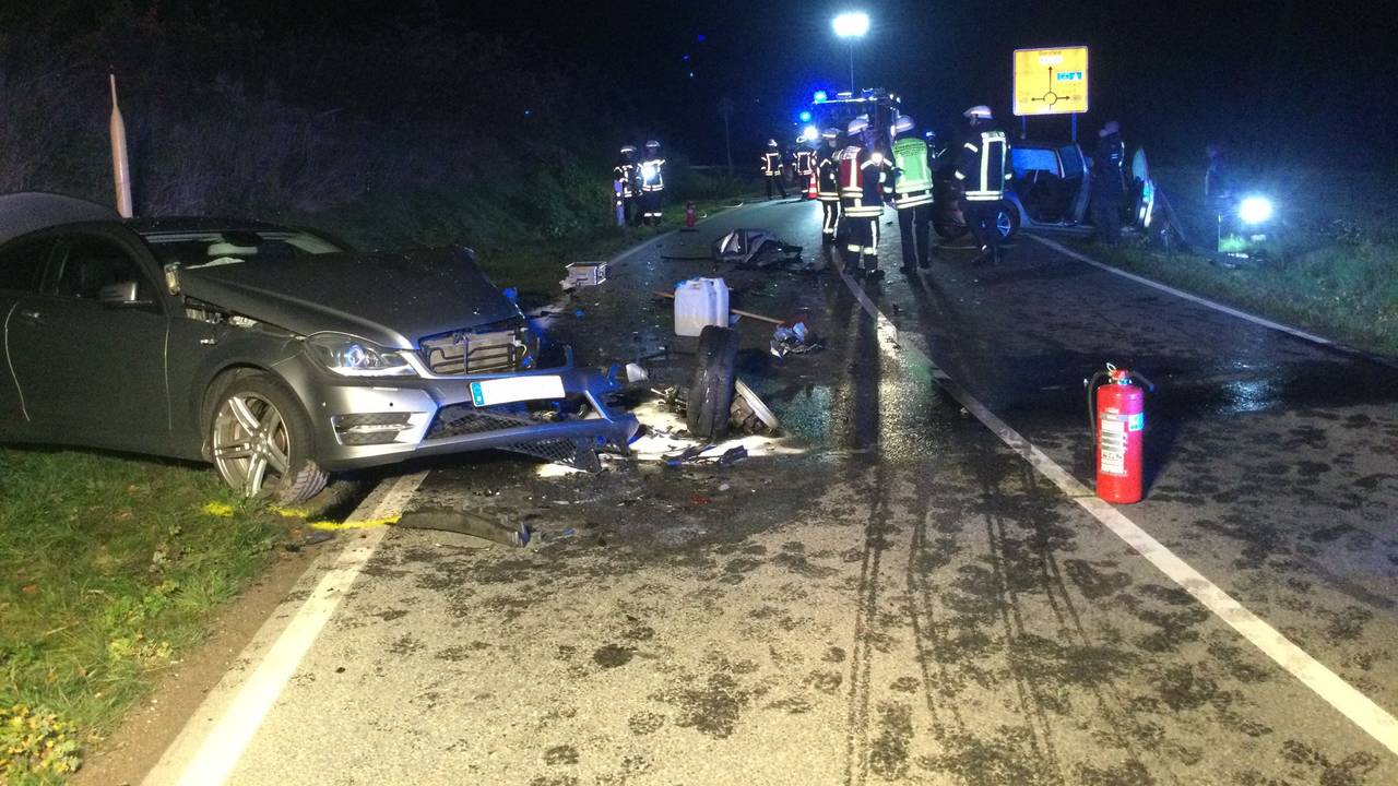 Ein beschädigtes Auto steht schräg neben der Straße, um es herum liegen Trümmerteile. Ein paar Meter weiter befinden sich mehrere Einsatzkräfte der Feuerwehr auf und neben der Fahrbahn.