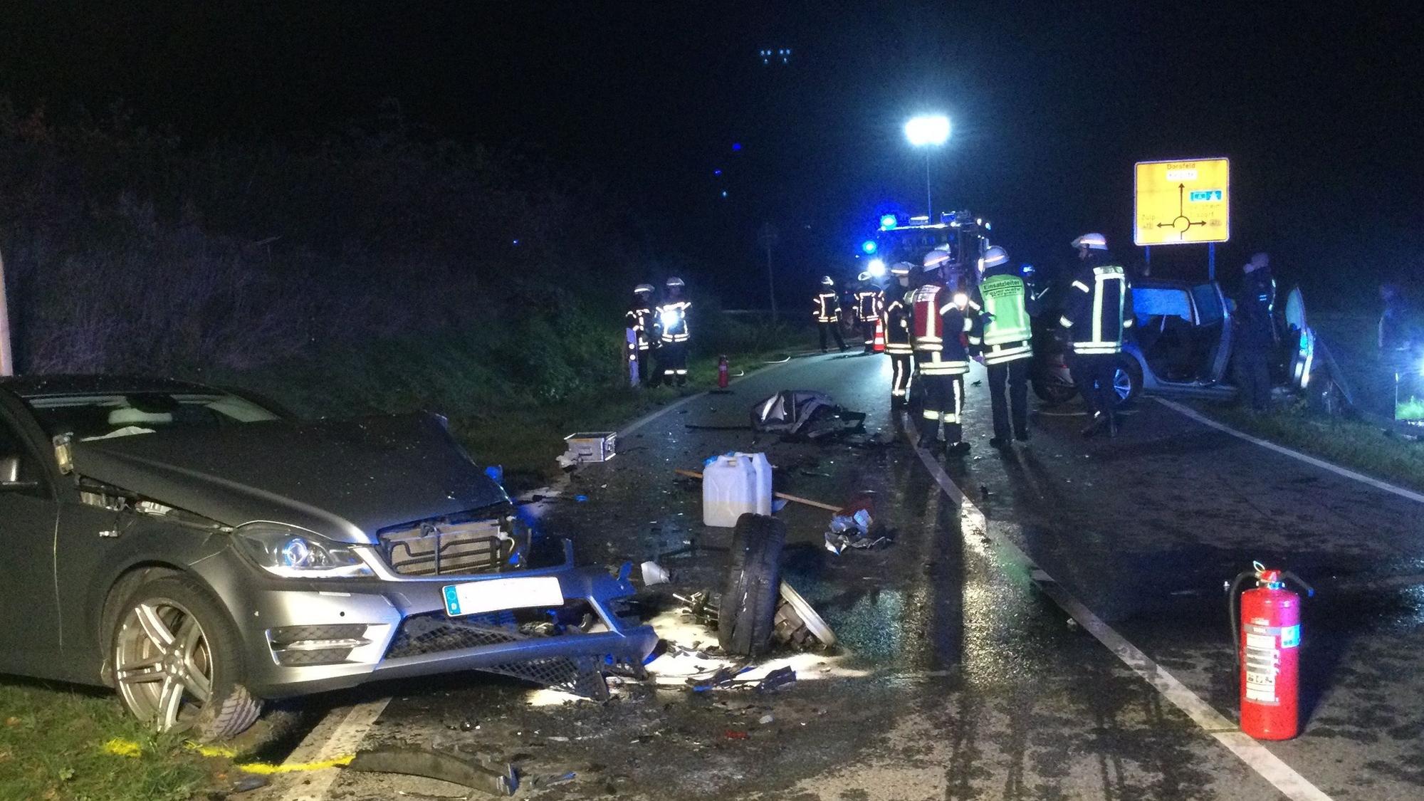 Ein beschädigtes Auto steht schräg neben der Straße, um es herum liegen Trümmerteile. Ein paar Meter weiter befinden sich mehrere Einsatzkräfte der Feuerwehr auf und neben der Fahrbahn.