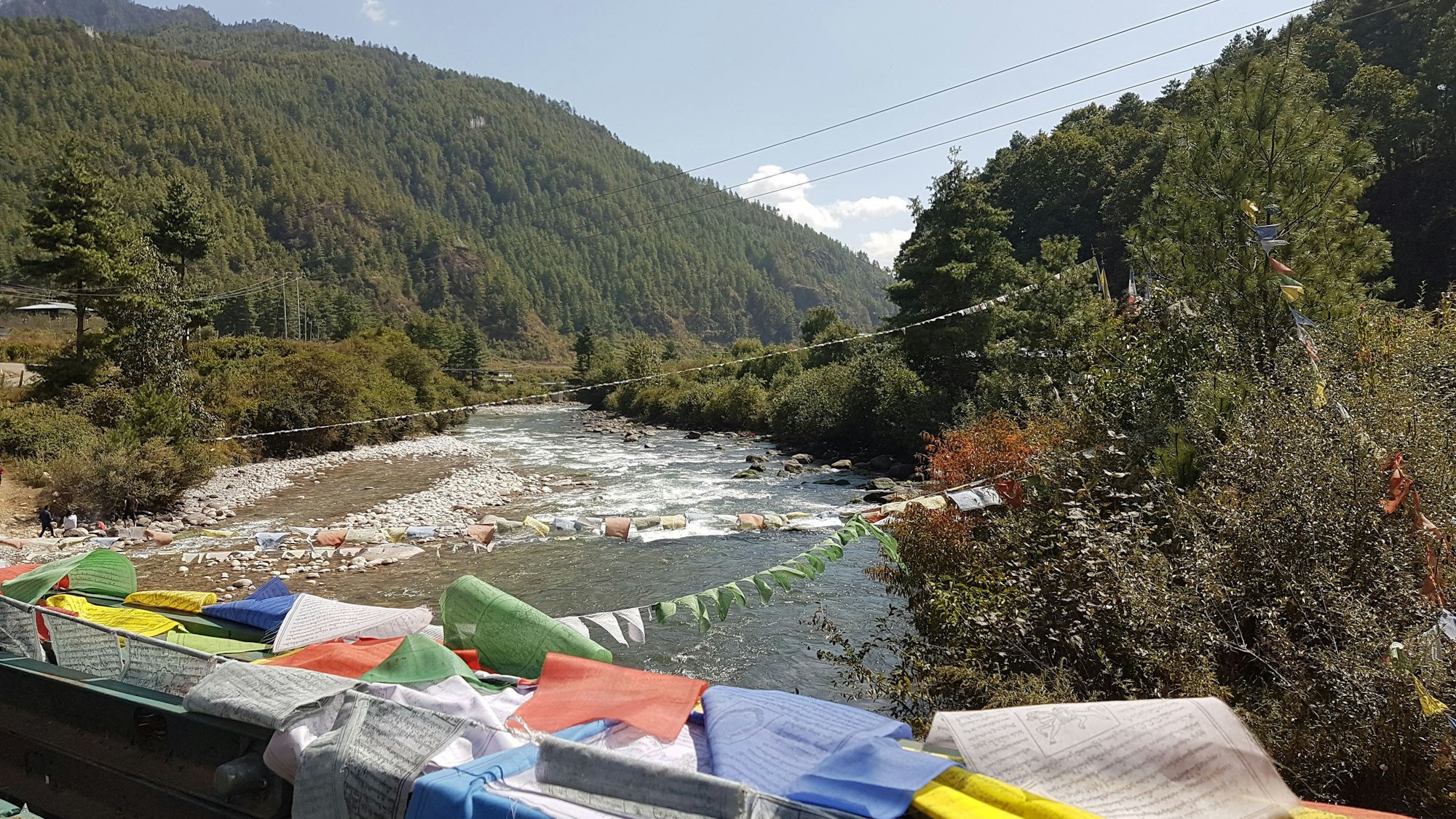 Gebetsfahnen und Stromleitungen kreuzen einen Fluss im Paro-Tal.