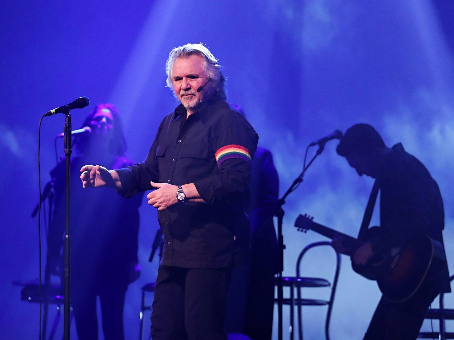 Tommy Engel steht mit schwarzem Hemd und Regenbogen-Binde am Arm auf der Bühne und singt.