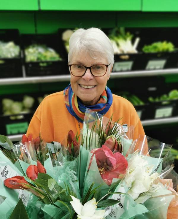 Ingrid Bauer steht vor einem Regal mit Gemüse. Sie hält einen Blumenstrauß in der Hand.