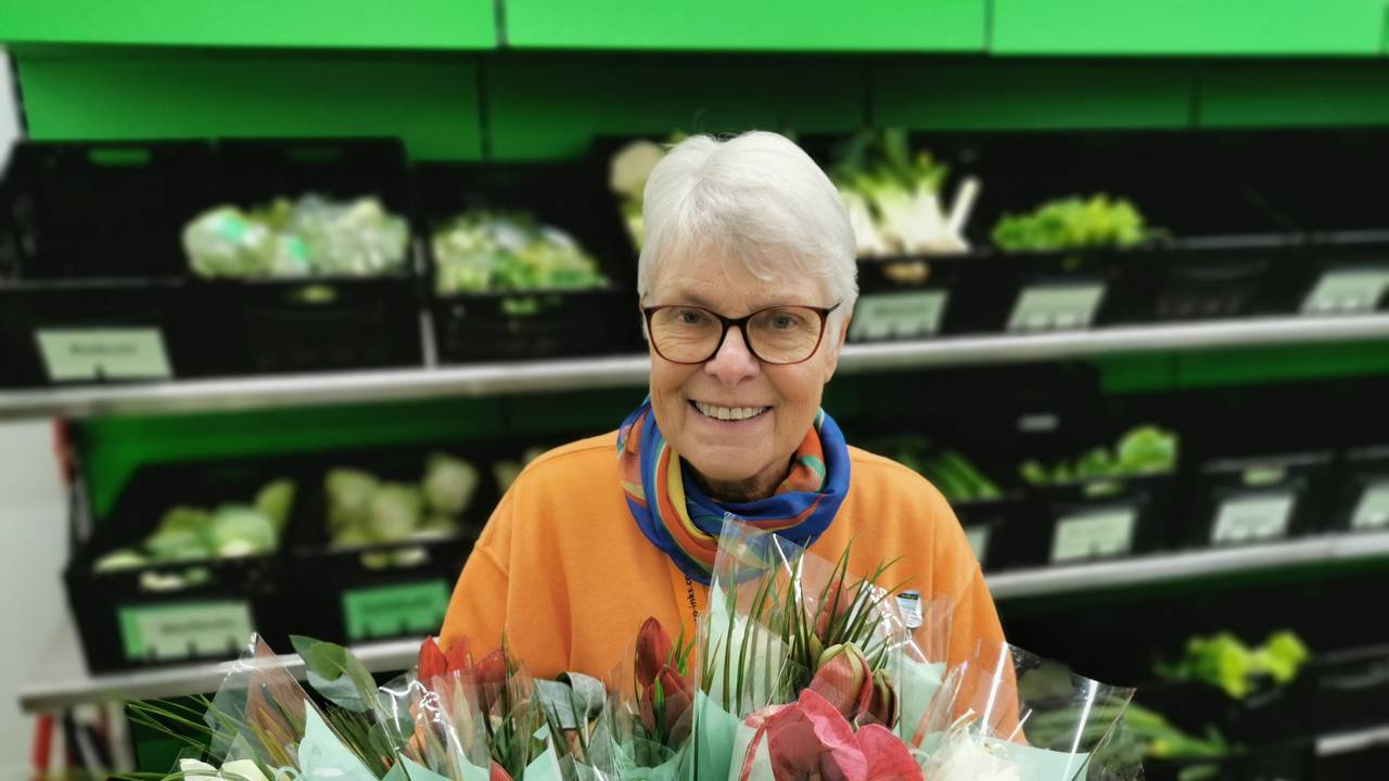 Ingrid Bauer steht vor einem Regal mit Gemüse. Sie hält einen Blumenstrauß in der Hand.