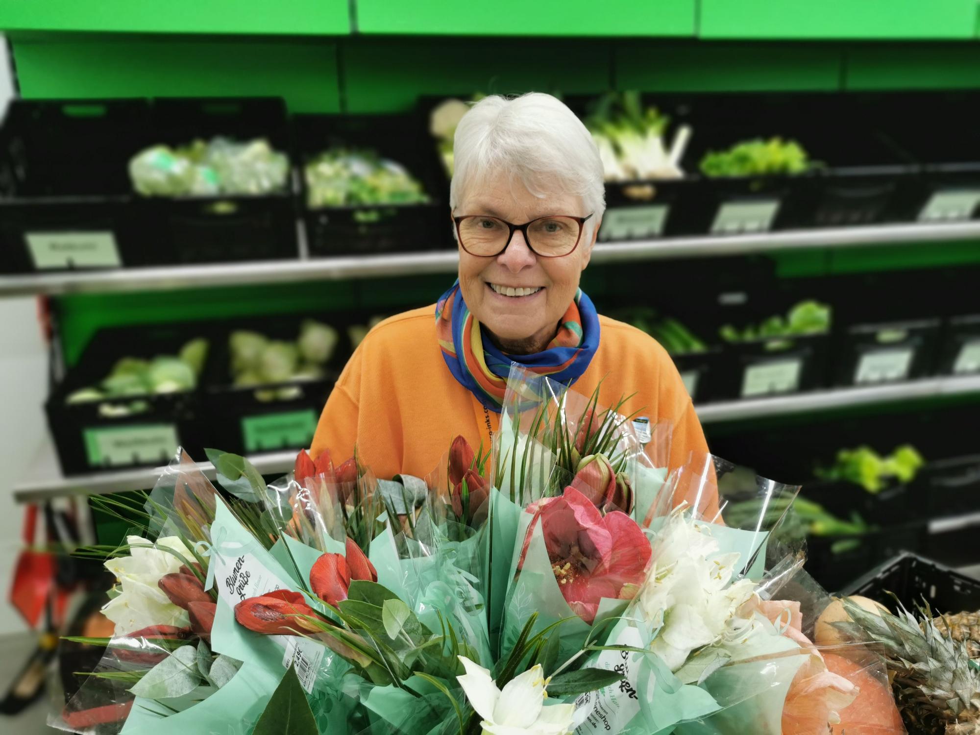 Ingrid Bauer steht vor einem Regal mit Gemüse. Sie hält einen Blumenstrauß in der Hand.