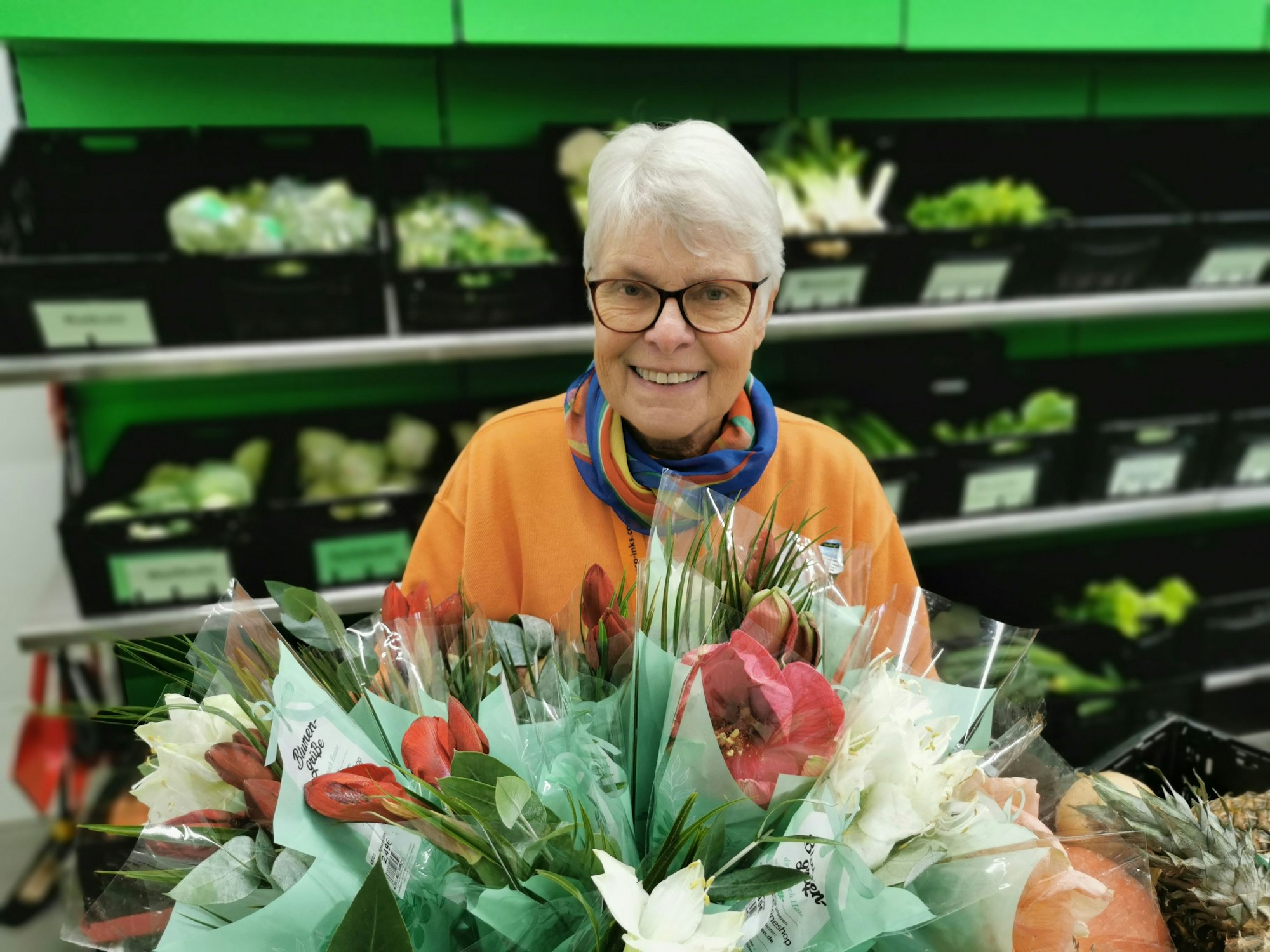 Ingrid Bauer steht vor einem Regal mit Gemüse. Sie hält einen Blumenstrauß in der Hand.