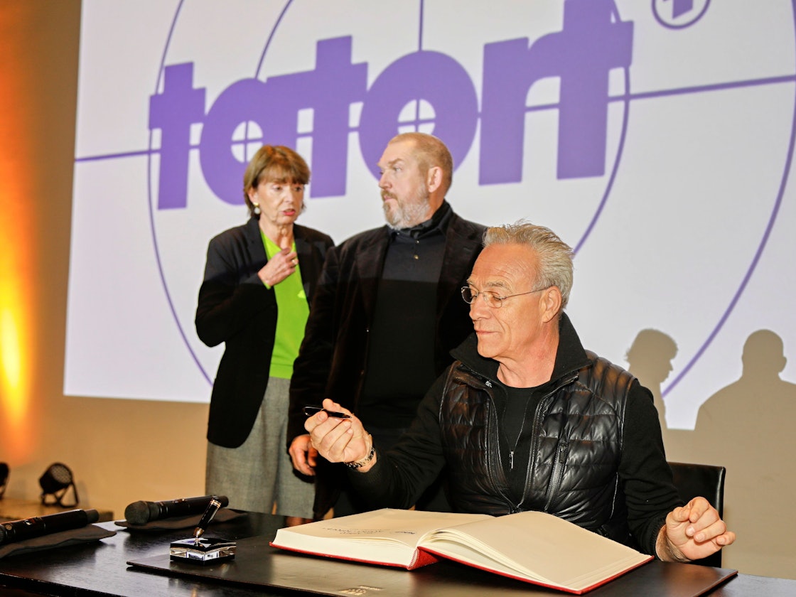 Die Tatort-Schauspieler Dietmar Bär und Klaus J. Behrendt tragen sich ins Gästebuch der Stadt Köln ein. Daneben steht OB Henriette Reker.