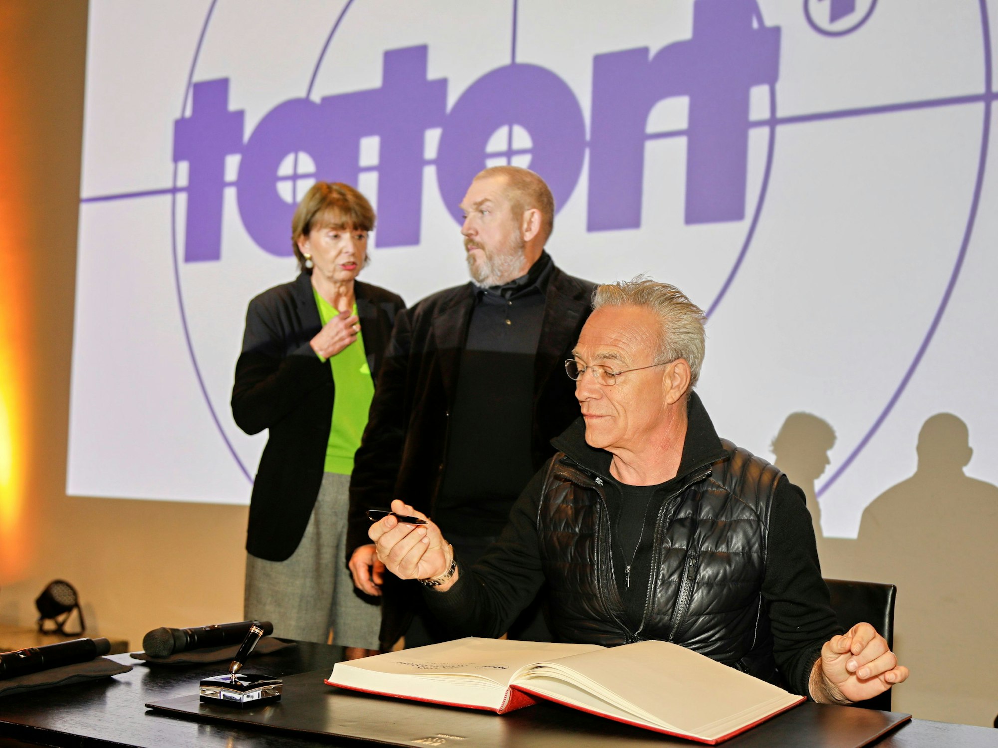 Die Tatort-Schauspieler Dietmar Bär und Klaus J. Behrendt tragen sich ins Gästebuch der Stadt Köln ein. Daneben steht OB Henriette Reker.