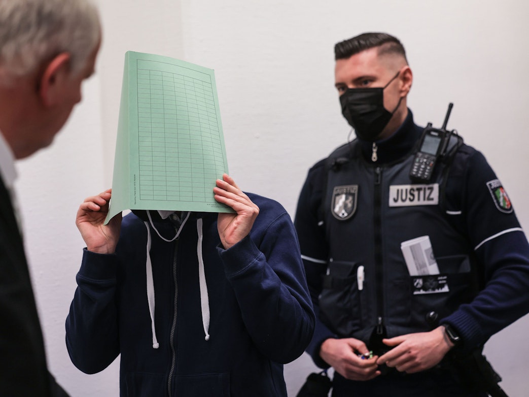 Ein Angeklagter hält sich vor Gericht einen Ordner vor das Gesicht. Neben ihm steht sein Verteidiger und ein Polizist.