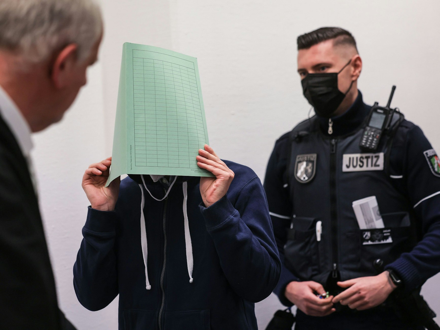 Ein Angeklagter hält sich vor Gericht einen Ordner vor das Gesicht. Neben ihm steht sein Verteidiger und ein Polizist.