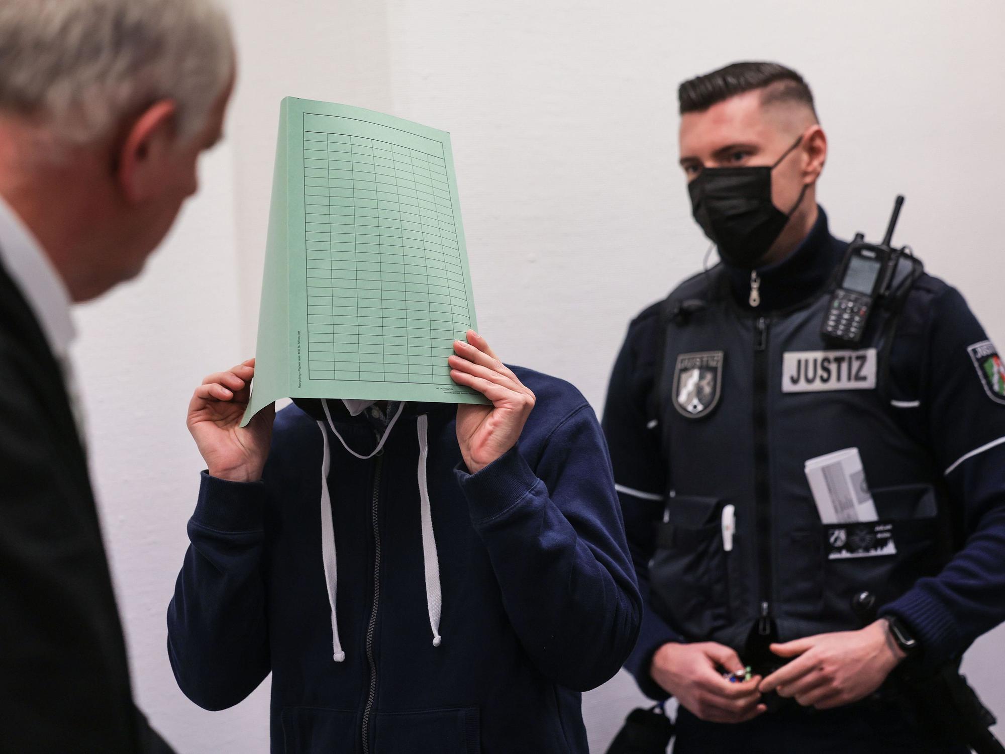 Ein Angeklagter hält sich vor Gericht einen Ordner vor das Gesicht. Neben ihm steht sein Verteidiger und ein Polizist.