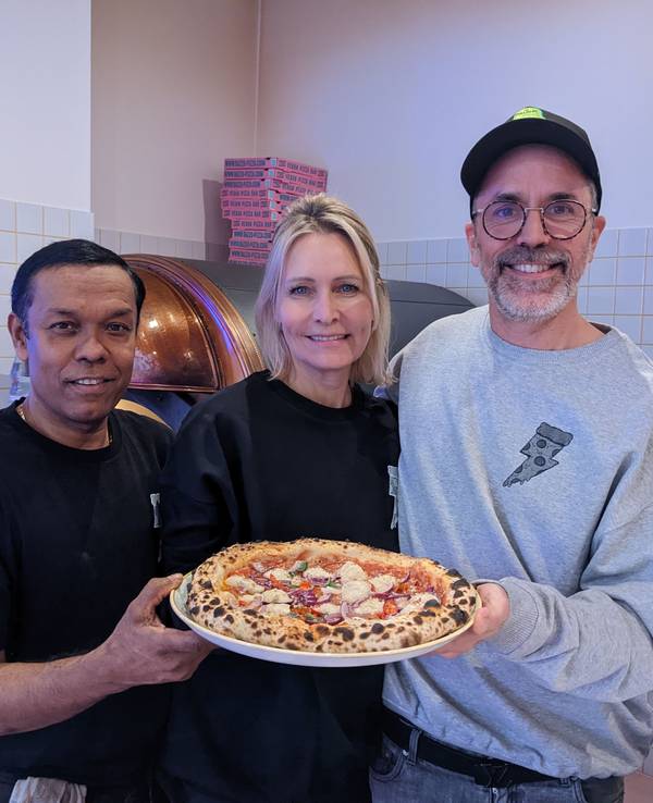 Pizzabäcker Monir (l.) posiert am 8. Dezember 2022 mit Chefin Verena Malta und Chef Ingo Noack. Sie halten einen Teller, auf dem sich eine vegane Pizza befindet.