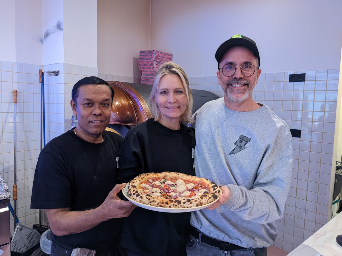 Pizzabäcker Monir (l.) posiert am 8. Dezember 2022 mit Chefin Verena Malta und Chef Ingo Noack. Sie halten einen Teller, auf dem sich eine vegane Pizza befindet.