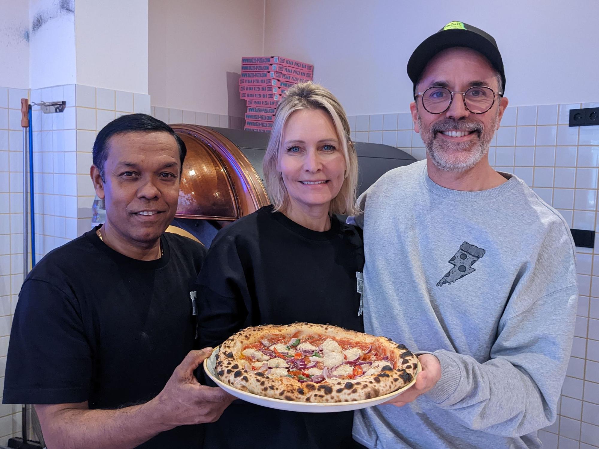 Pizzabäcker Monir (l.) posiert am 8. Dezember 2022 mit Chefin Verena Malta und Chef Ingo Noack. Sie halten einen Teller, auf dem sich eine vegane Pizza befindet.
