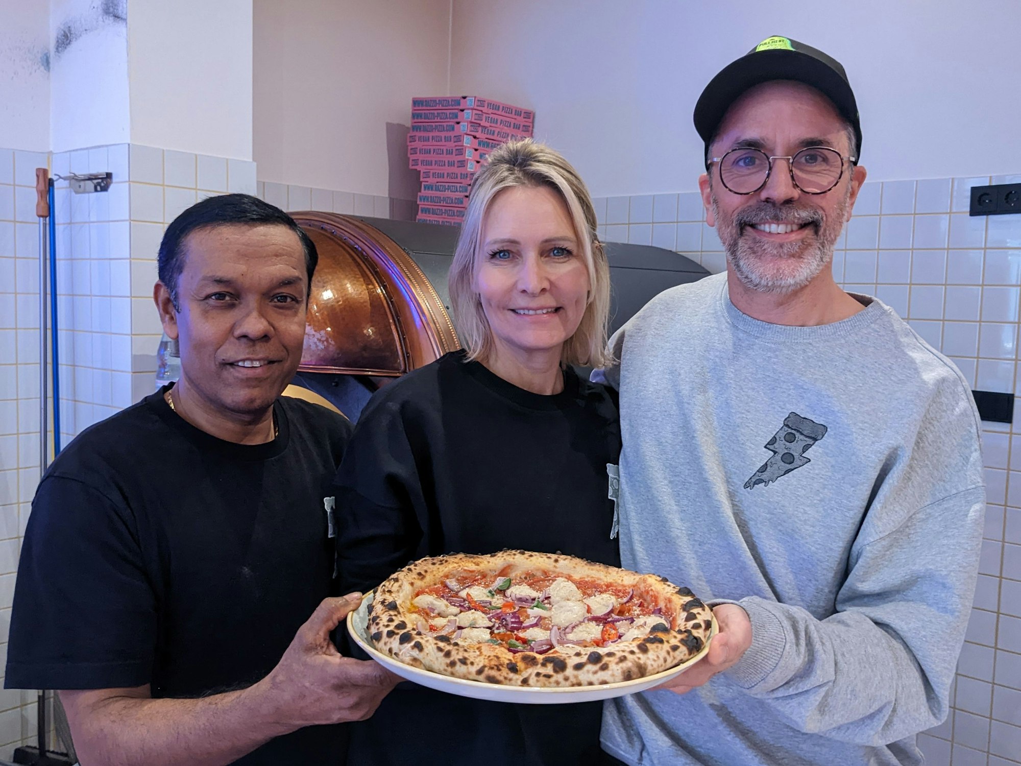 Pizzabäcker Monir (l.) posiert am 8. Dezember 2022 mit Chefin Verena Malta und Chef Ingo Noack. Sie halten einen Teller, auf dem sich eine vegane Pizza befindet.