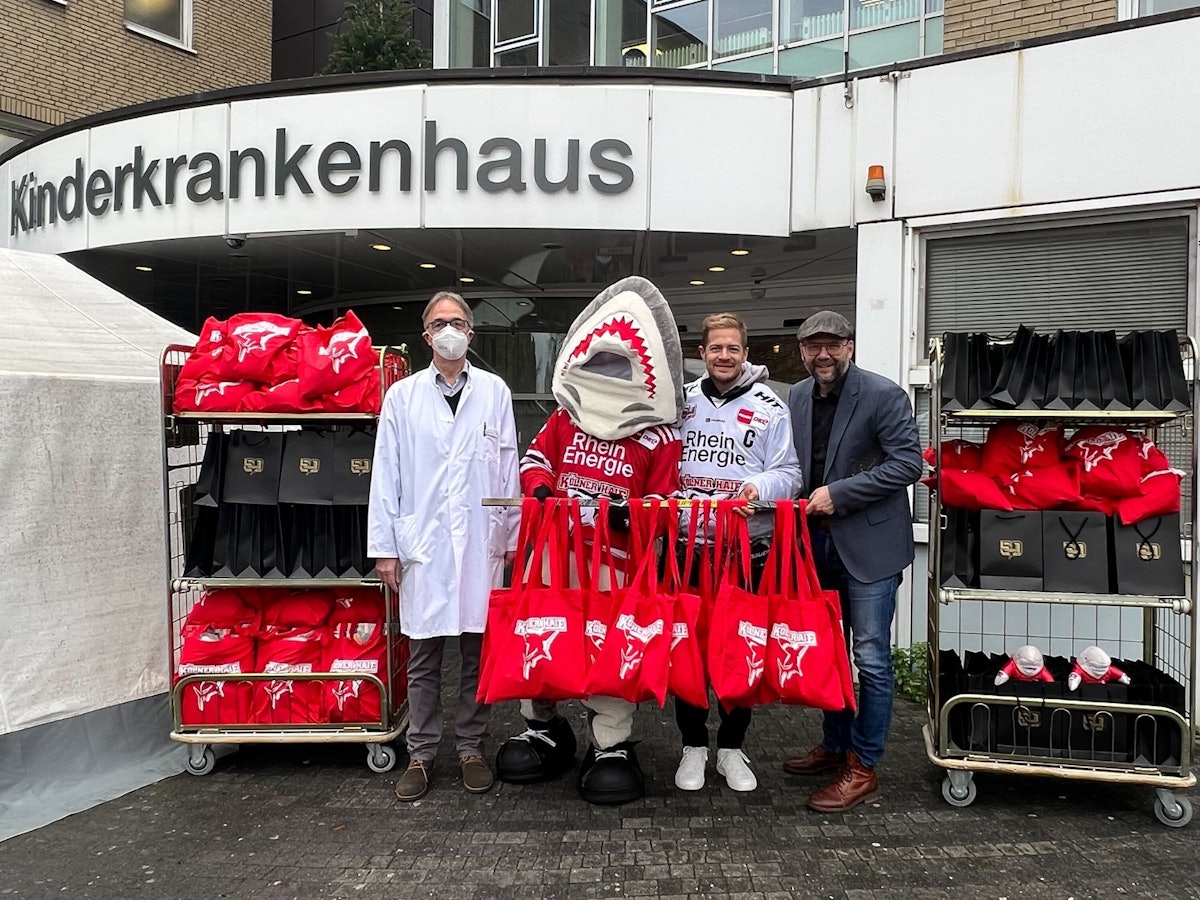 Professor Michael Weiß, Sharky, KEC-Kapitän Moritz Müller sowie Haie-Geschäftsführer Philipp Walter mit den Geschenken vor der Kinderklinik.