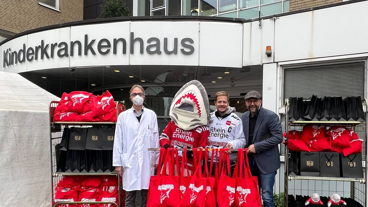 Professor Michael Weiß, Sharky, KEC-Kapitän Moritz Müller sowie Haie-Geschäftsführer Philipp Walter mit den Geschenken vor der Kinderklinik.
