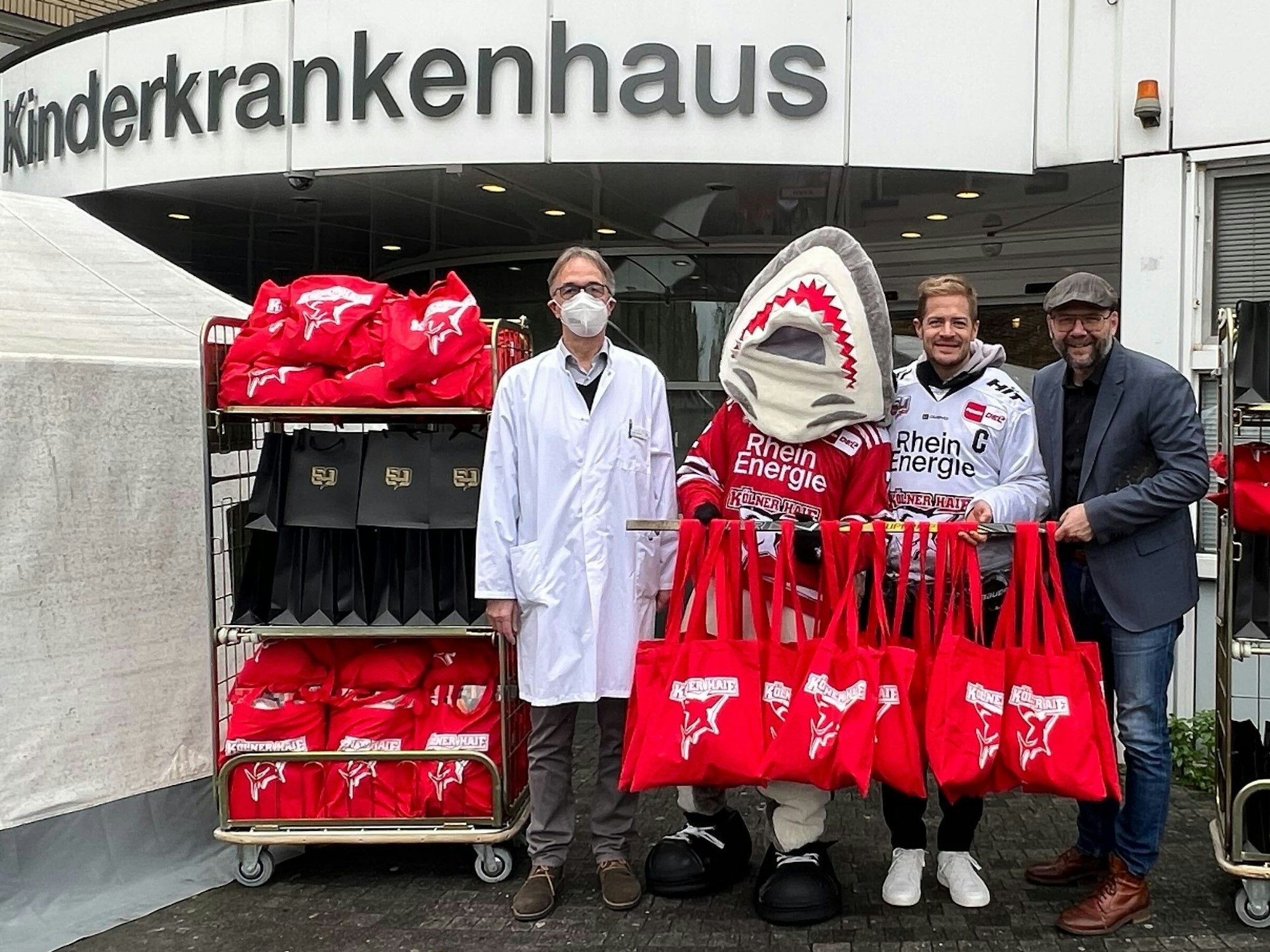 Professor Michael Weiß, Sharky, KEC-Kapitän Moritz Müller sowie Haie-Geschäftsführer Philipp Walter mit den Geschenken vor der Kinderklinik.