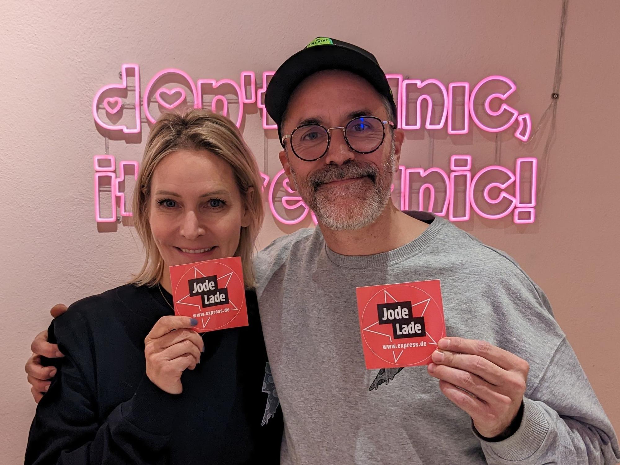 Verena Malta und Ingo Noack vom Razzo halten „Jode Lade“-Sticker hoch. Diese Auszeichnung haben sie von EXPRESS.de von erhalten. Im Hintergrund ist der Slogan „Don't panic, it's veganic“ zu lesen.