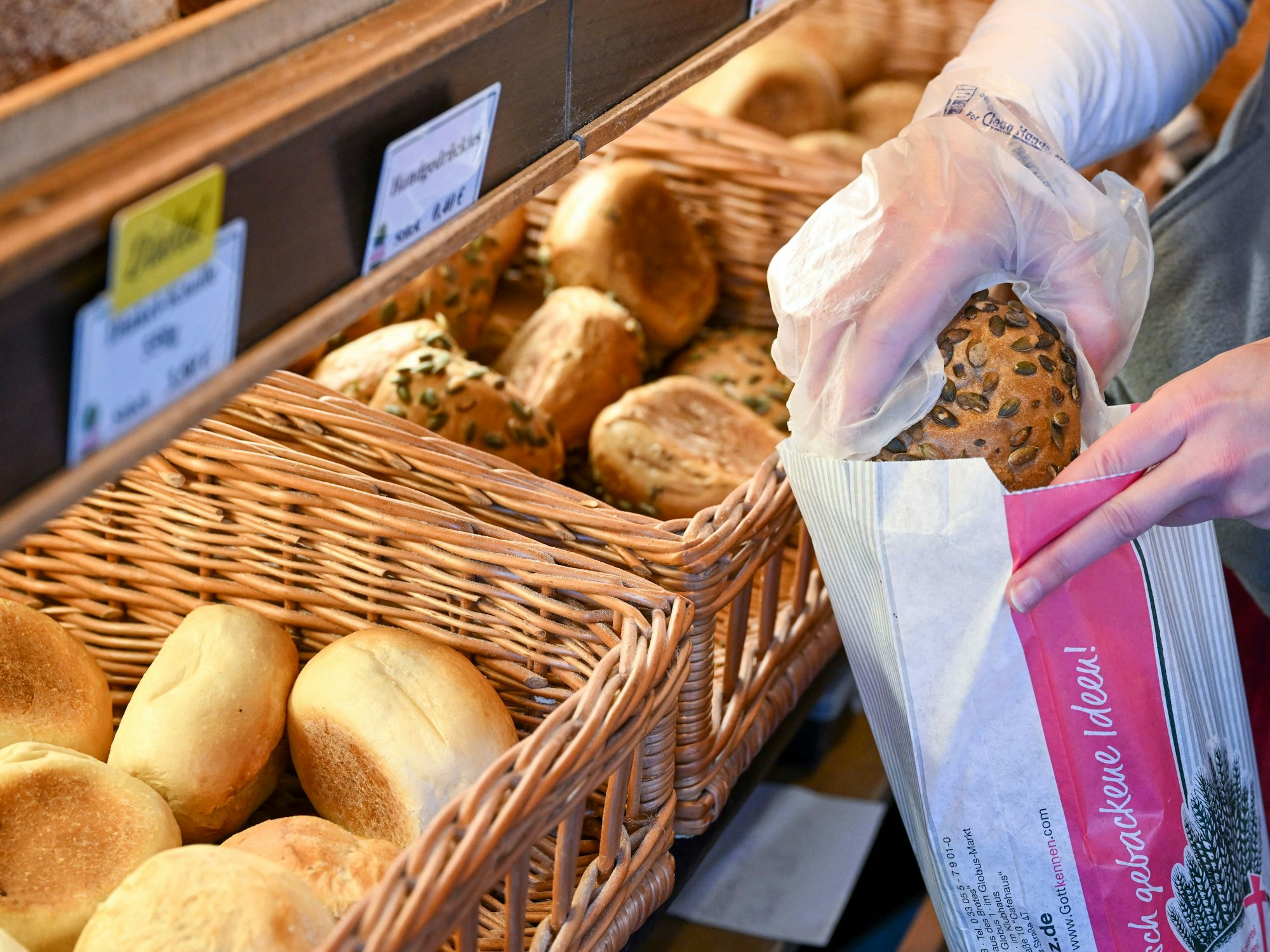 Eine Person packt ein Körnerbrötchen in eine Papiertüte in einer Bäckerei.